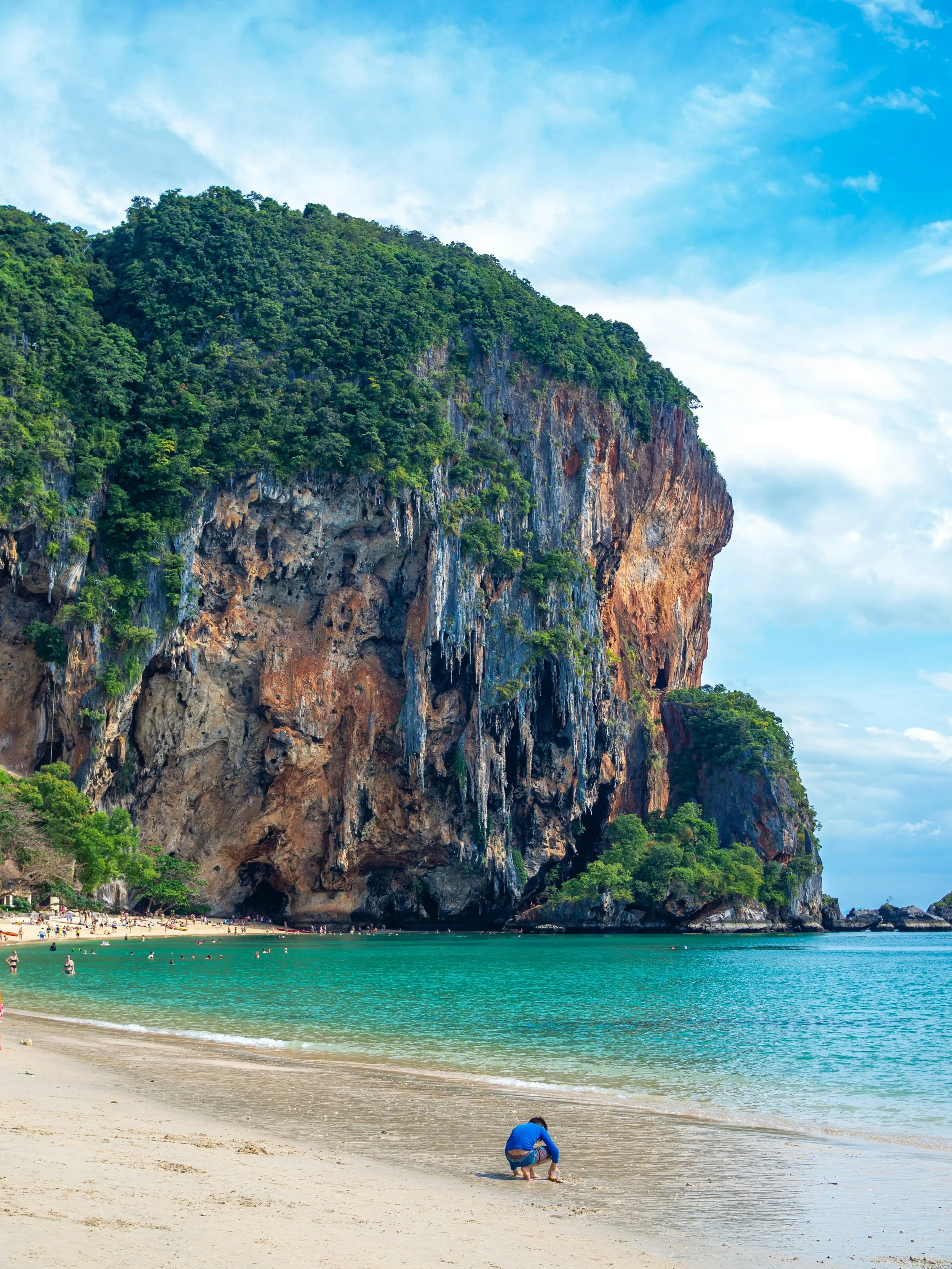 A tropical beach with a large rocky cliff covered in greenery, turquoise water, and a sandy shore, with people enjoying the beach and a person crouching near the water.