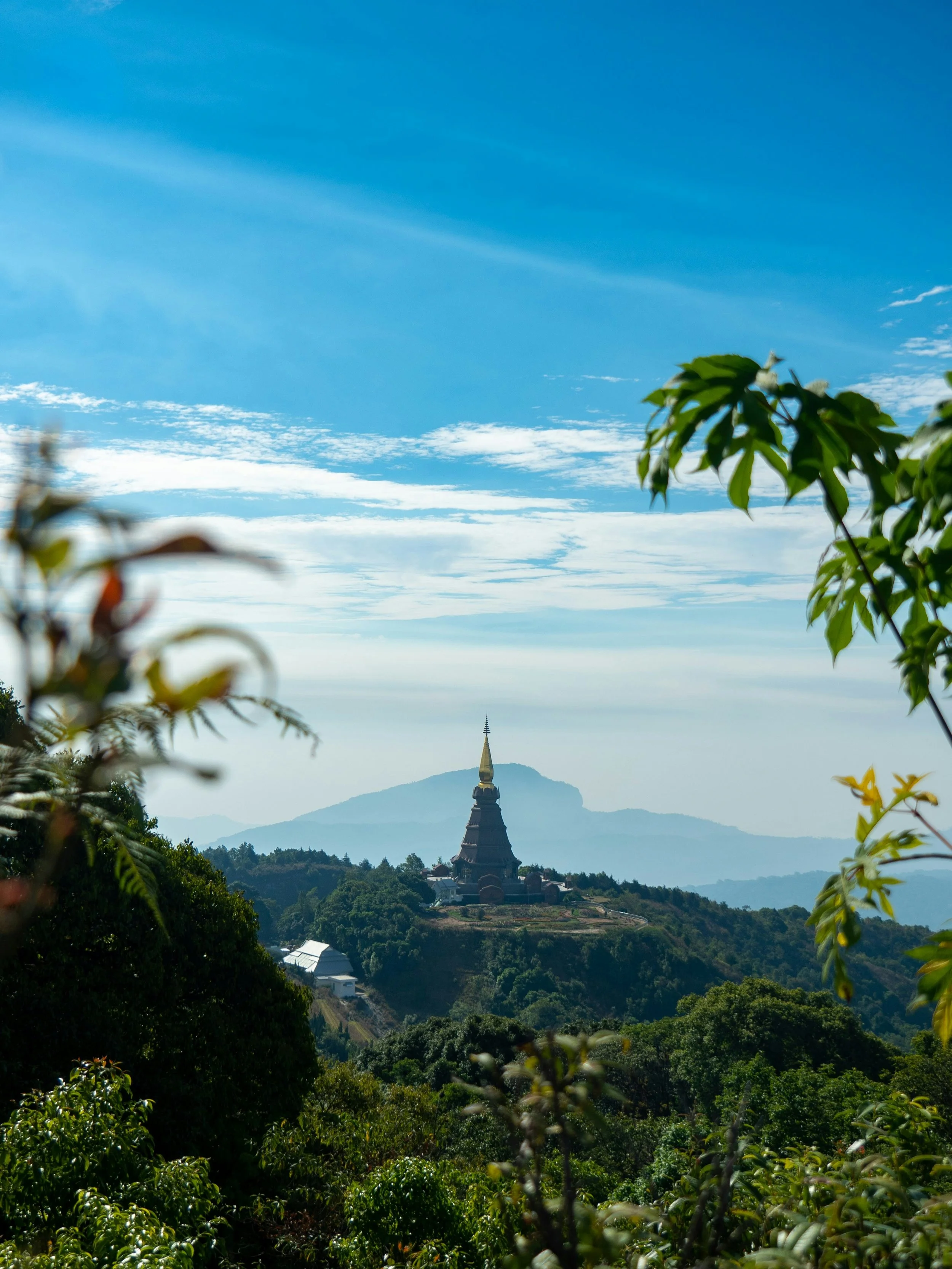 Scenic view of a hilltop temple with a golden spire, surrounded by lush green trees, with a mountain and blue sky in the background.