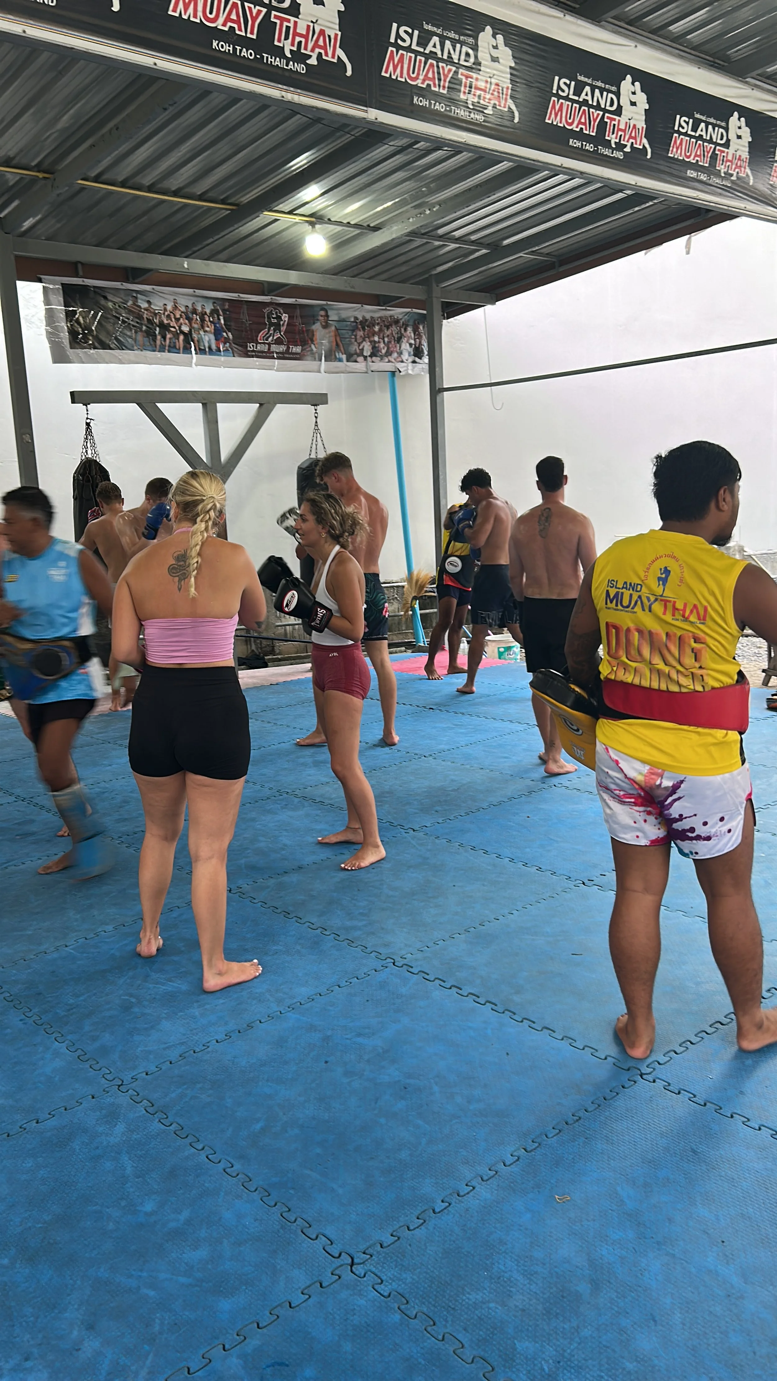 People practicing Muay Thai in a gym with blue mats, some wearing gloves, in Koh Tao, Thailand.