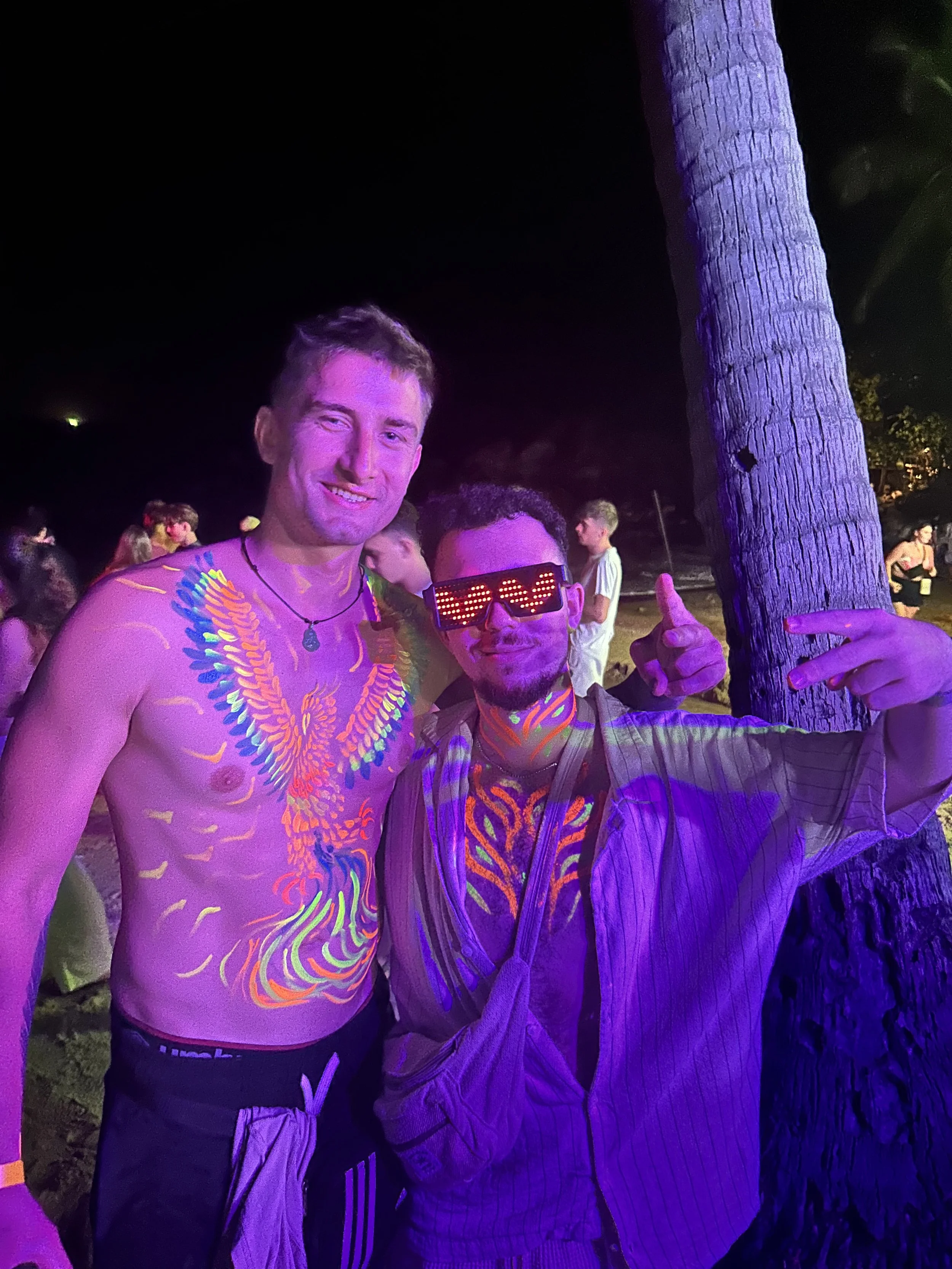 Two men standing outdoors at night, illuminated with blacklight, with neon body paint and one wearing glowing LED glasses. One man is shirtless with neon paint on his chest and a necklace, while the other wears a striped shirt, has neon paint on his chest, and is pointing at the camera.