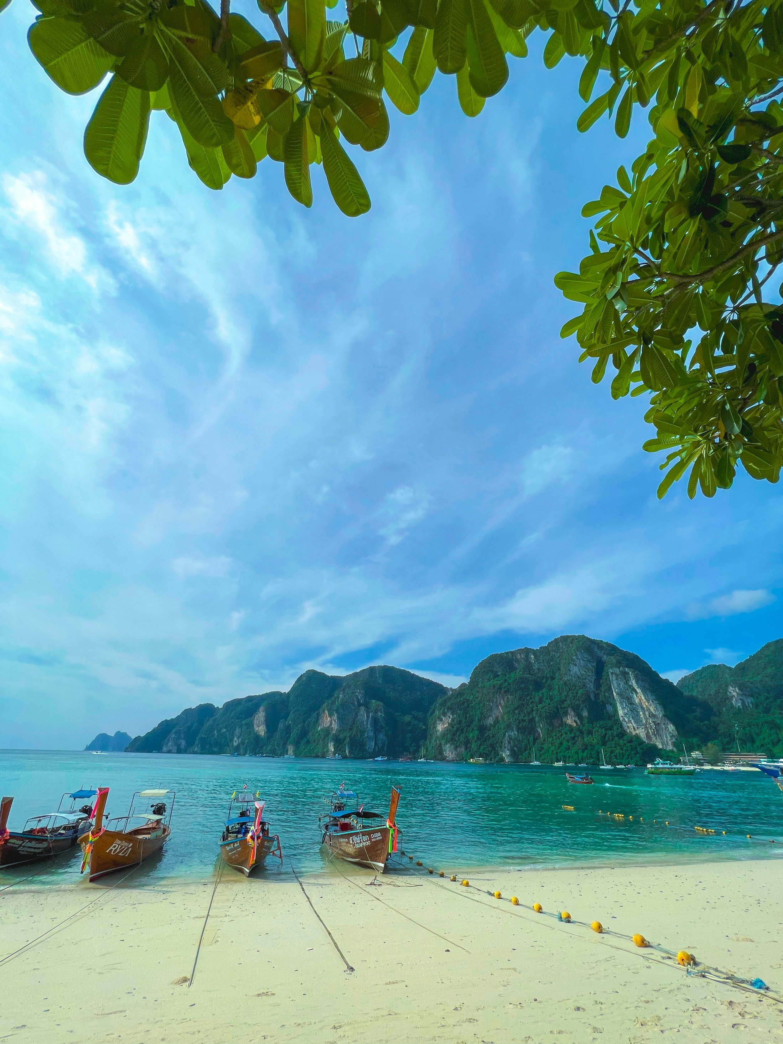 A beach scene with several boats anchored on the sandy shore, turquoise water, lush green mountains in the background, and a blue sky with some clouds overhead.