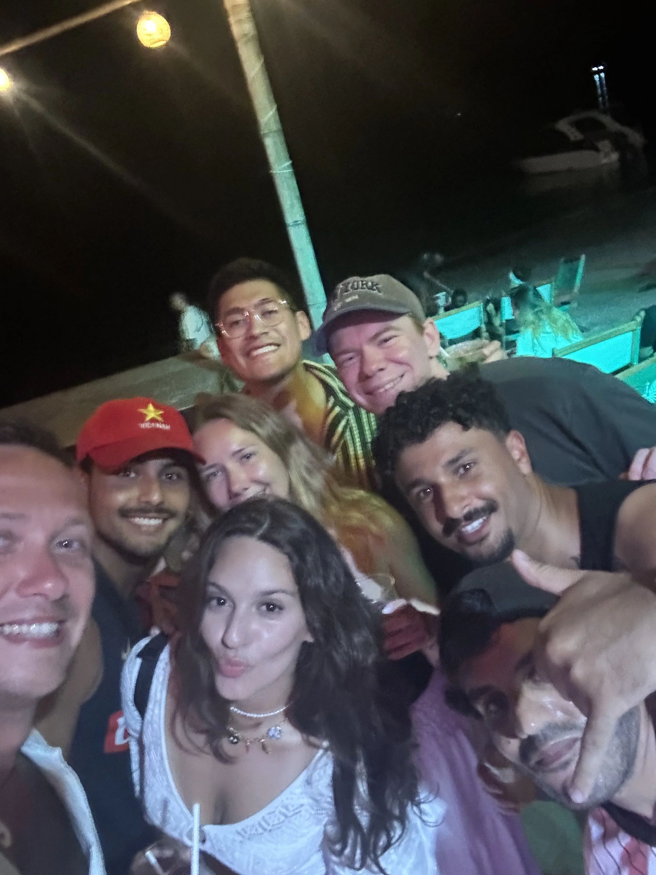 Group of nine people smiling and posing for a photo at night, with a boat and water in the background.
