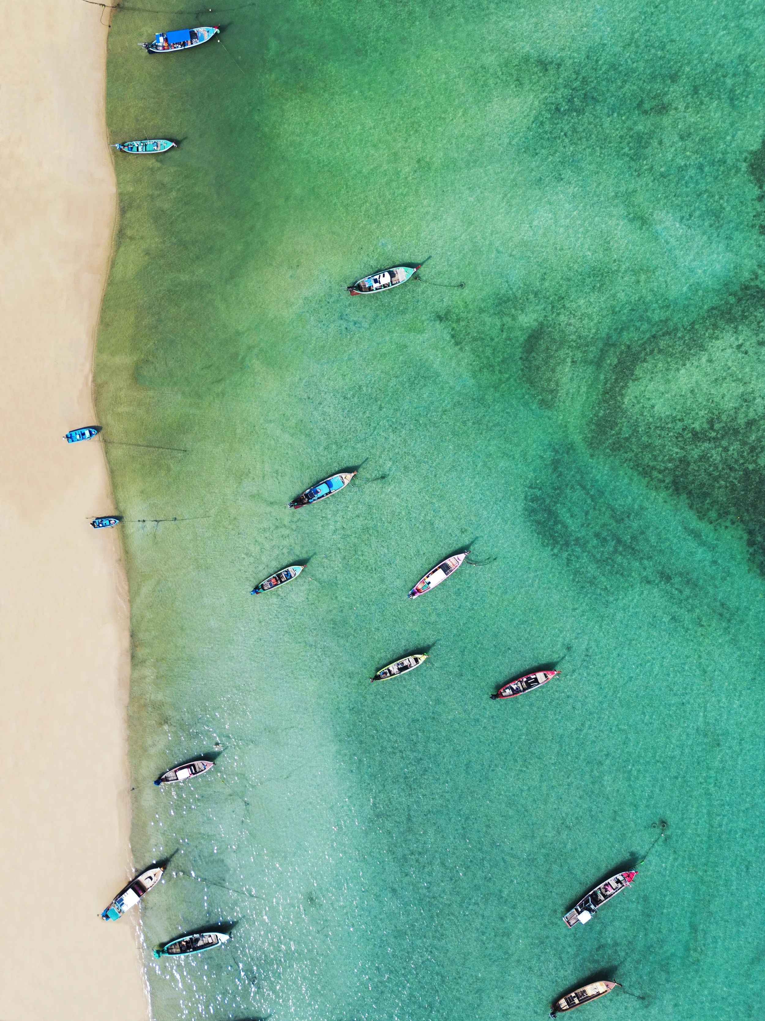 Aerial view of boats anchored near a sandy beach, with clear green water and seabed visible.