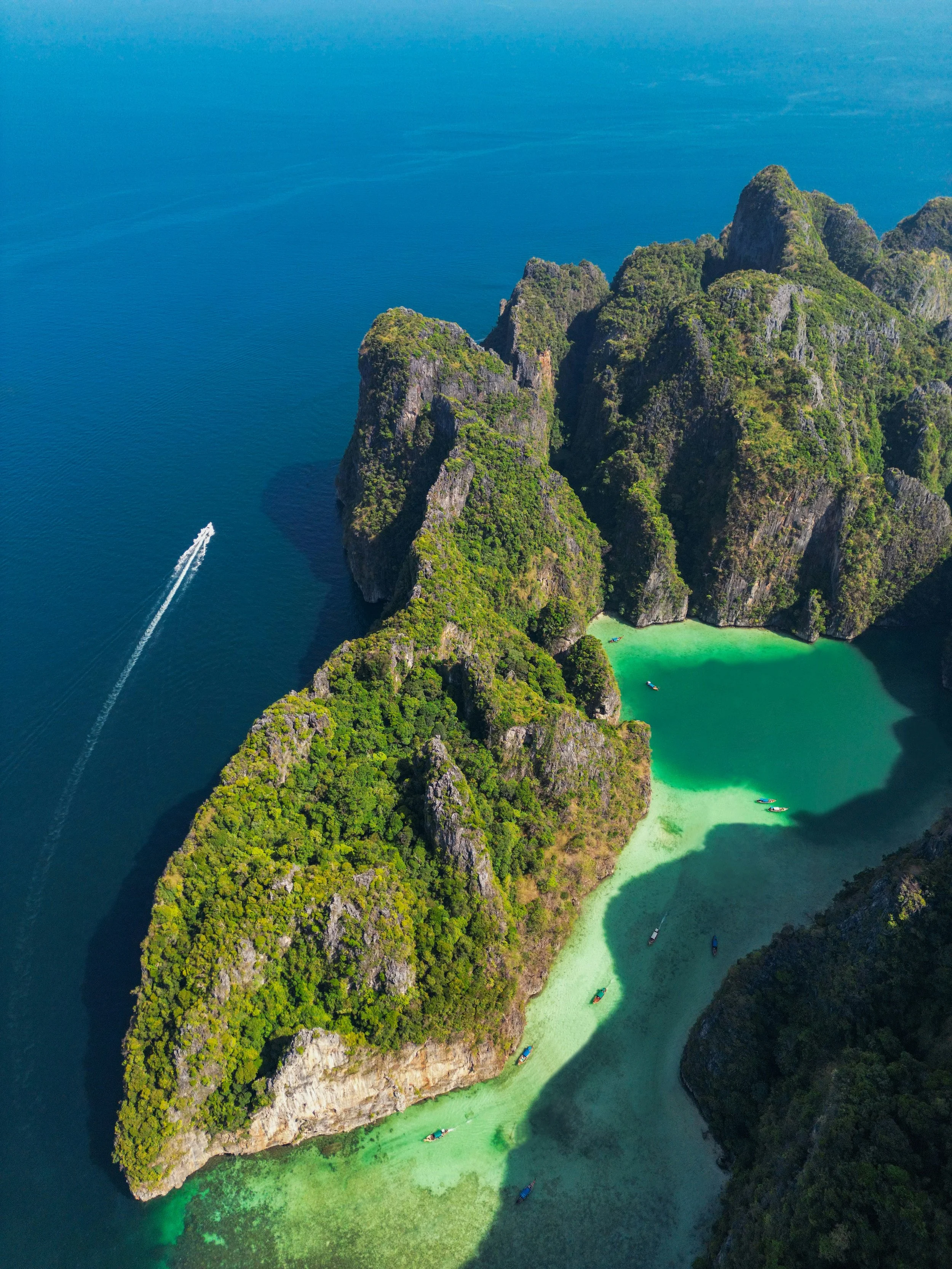 Aerial view of limestone cliffs covered with greenery surrounding a small, turquoise lagoon with boats; blue ocean with a boat leaving a white wake.