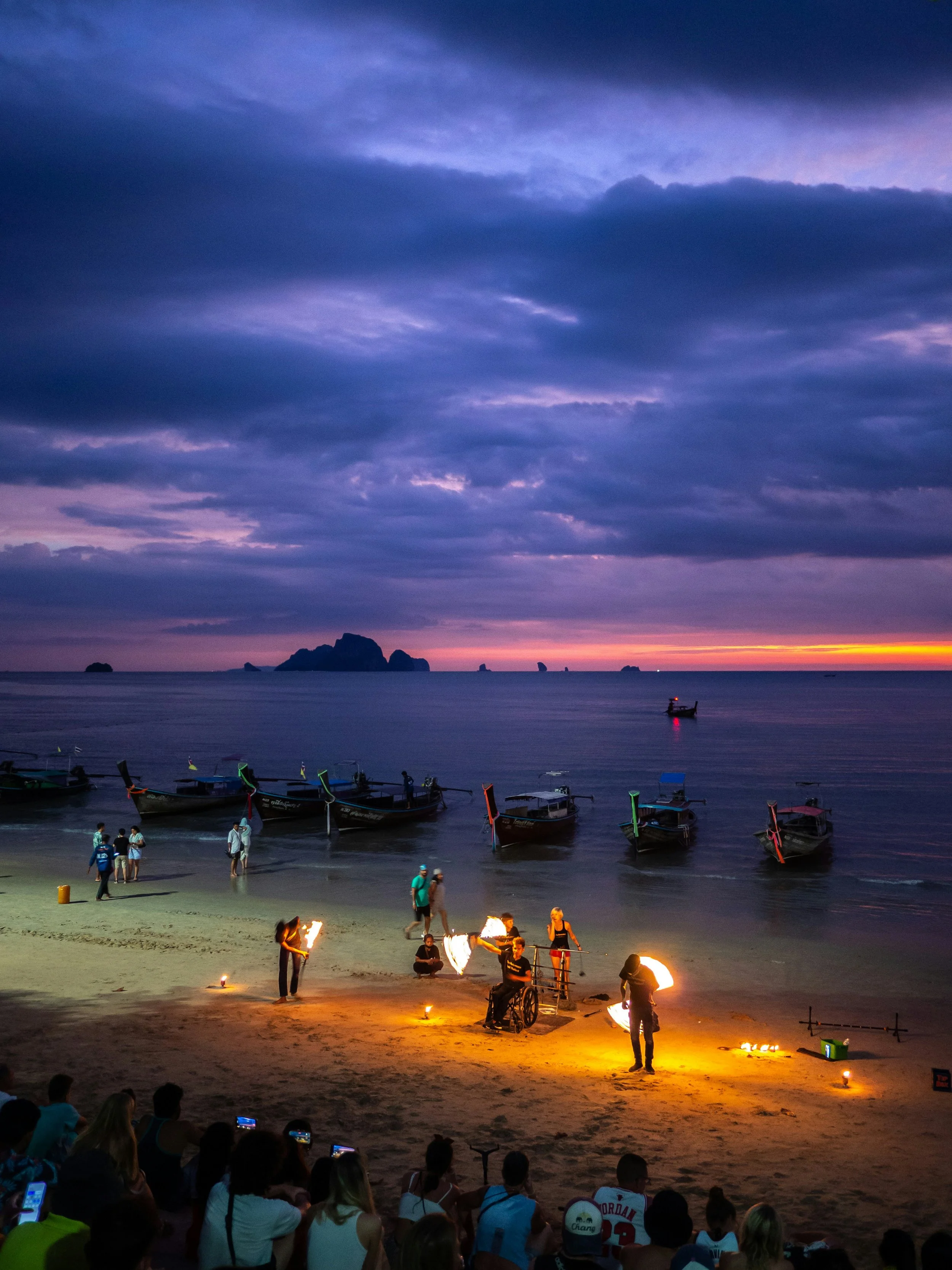 A beach scene during sunset with a fire show performed by fire performers, an audience seated nearby, boats anchored in the water, and a rocky island in the background under a partly cloudy sky.