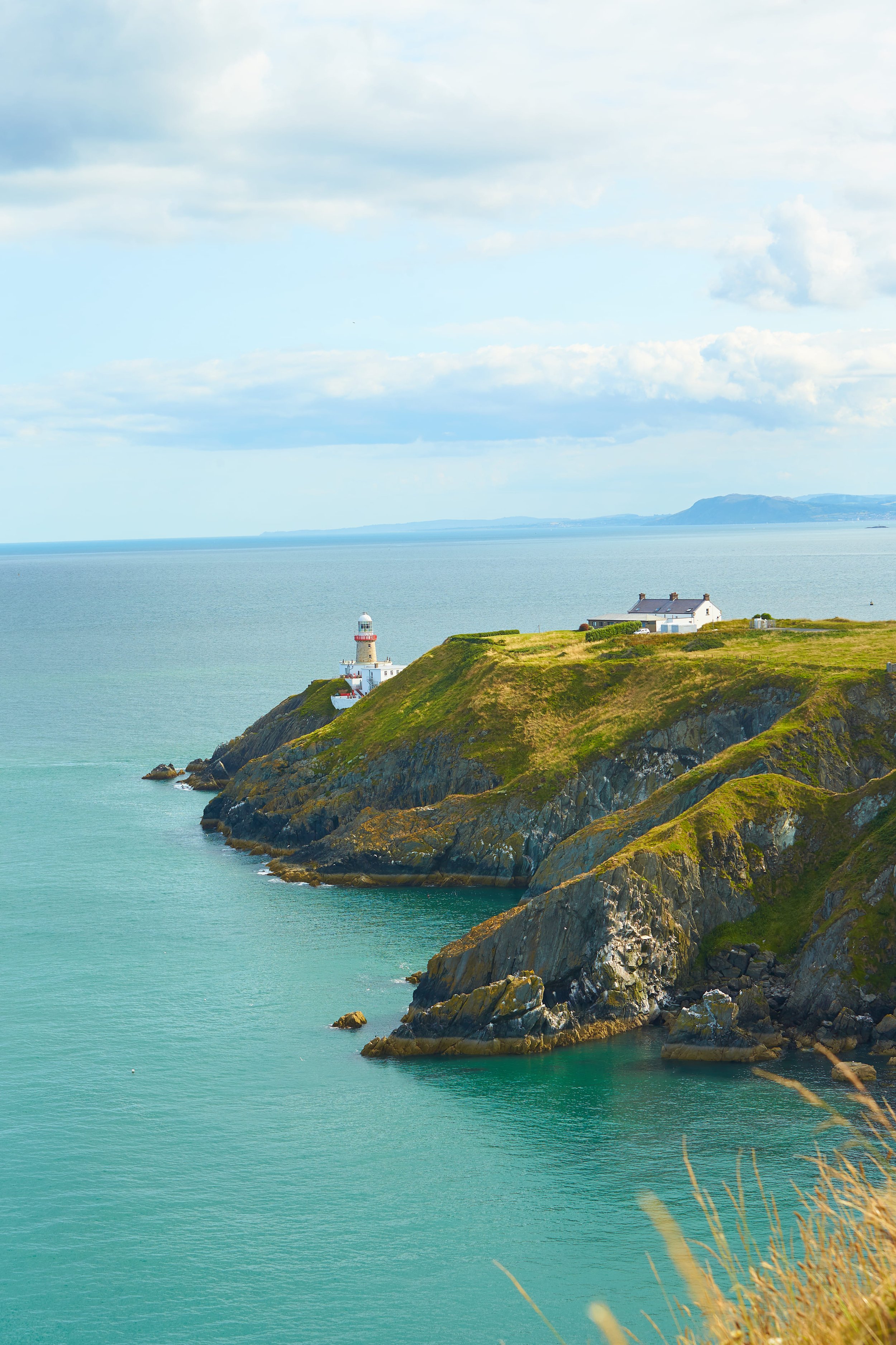 Scenic view of a lighthouse on a rocky green coastline overlooking the sea, with a house nearby and mountains in the distance under a partly cloudy sky.