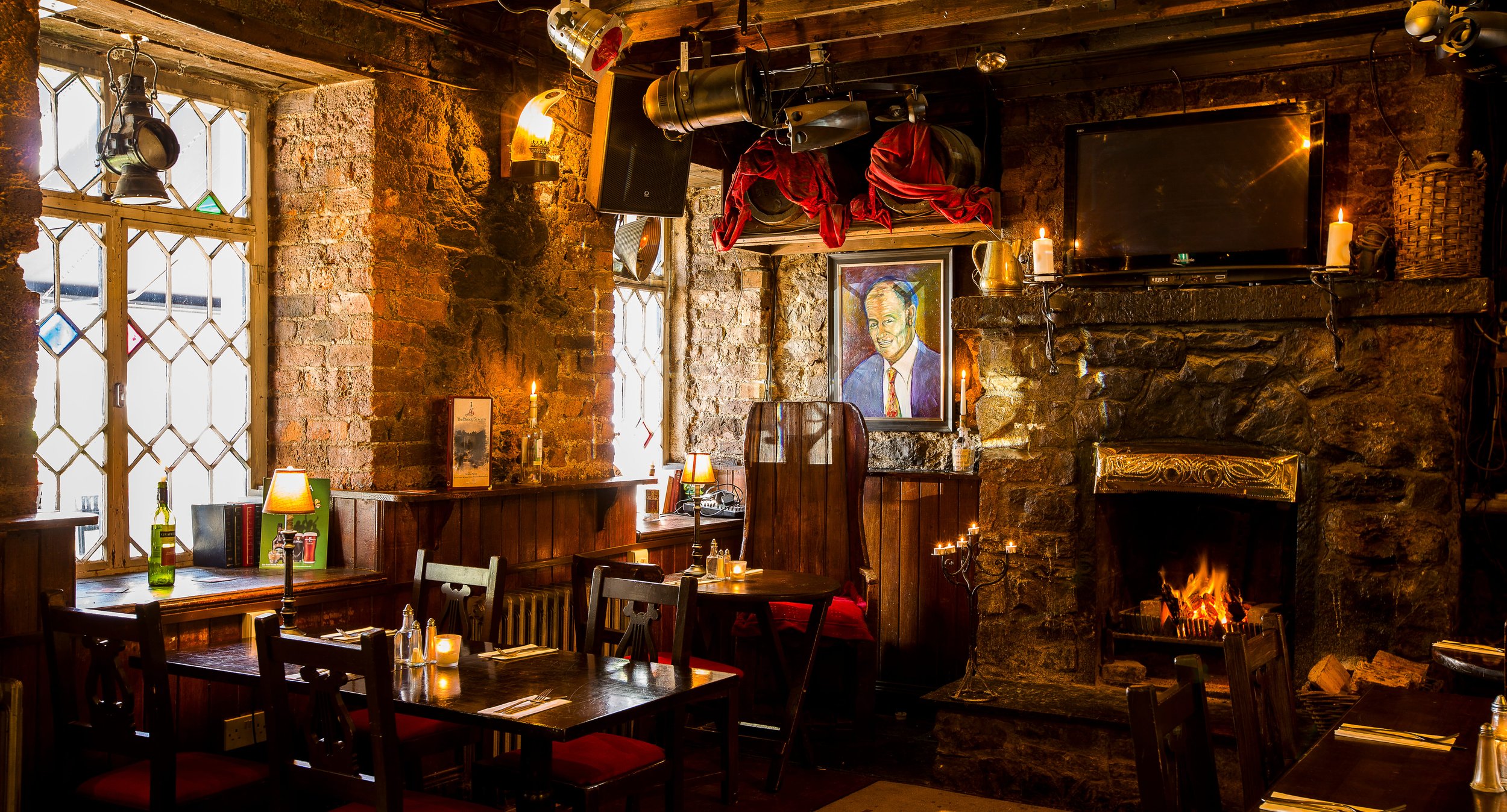 Cozy restaurant interior with brick walls, a fireplace with a lit fire, wooden furniture, candles, a painting of a man, a TV, and windows with diamond-pane glass.