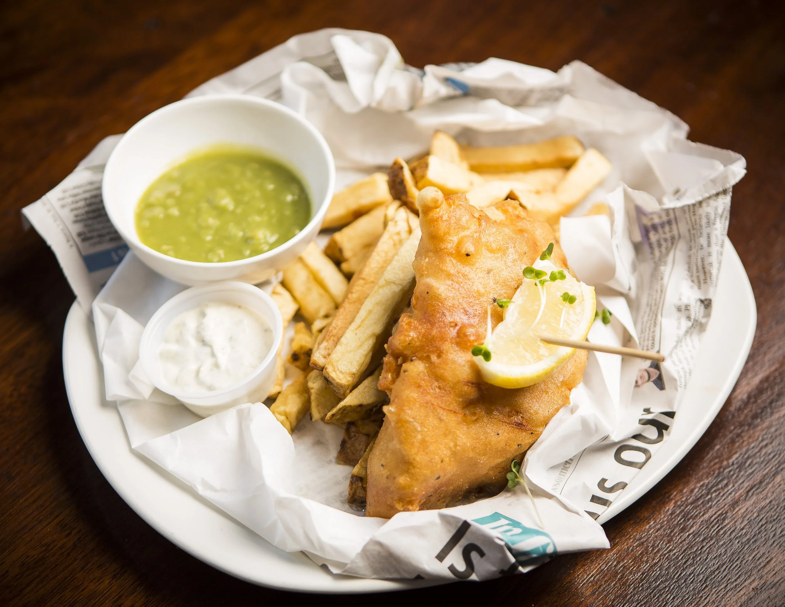Fried fish fillet with lemon wedge, served with French fries, tartar sauce, and green dipping sauce on a white plate lined with paper, on a dark wooden table.