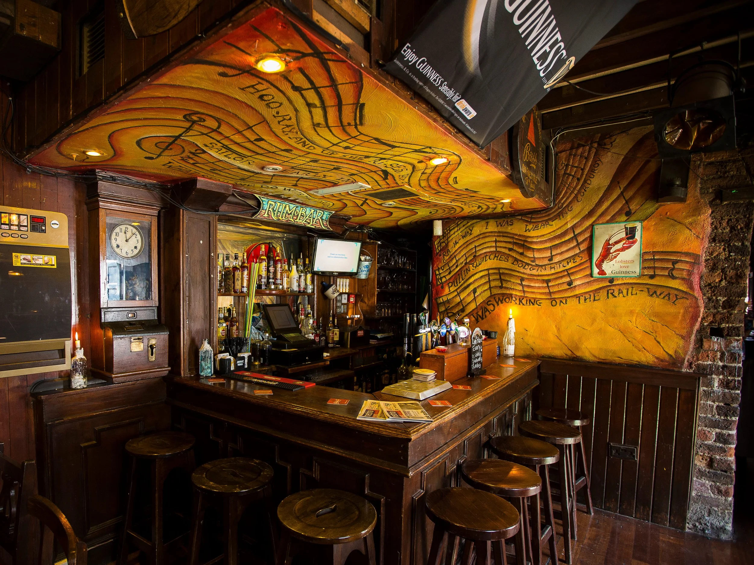 Inside a cozy bar with a wooden counter, barstools, a colorful mural with musical notes, and alcohol bottles behind the bar.