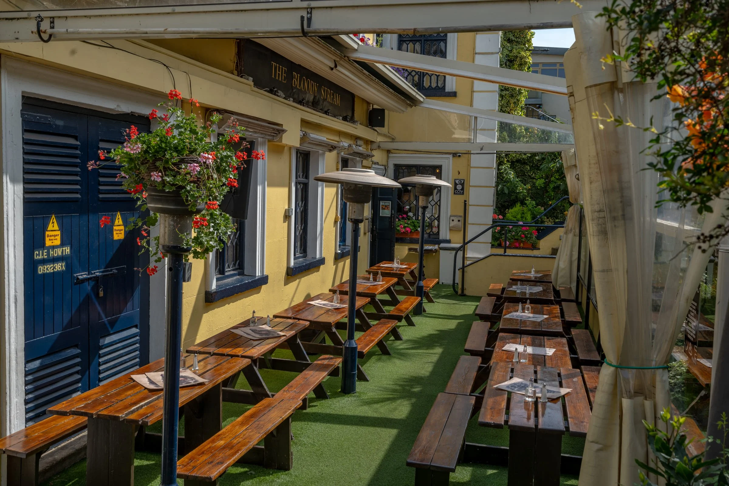 An outdoor patio area with wooden picnic tables set with napkins and glassware, alongside patio heaters and hanging flower baskets, adjacent to a yellow building with blue window frames and a sign reading 'The Bloody Stream.'