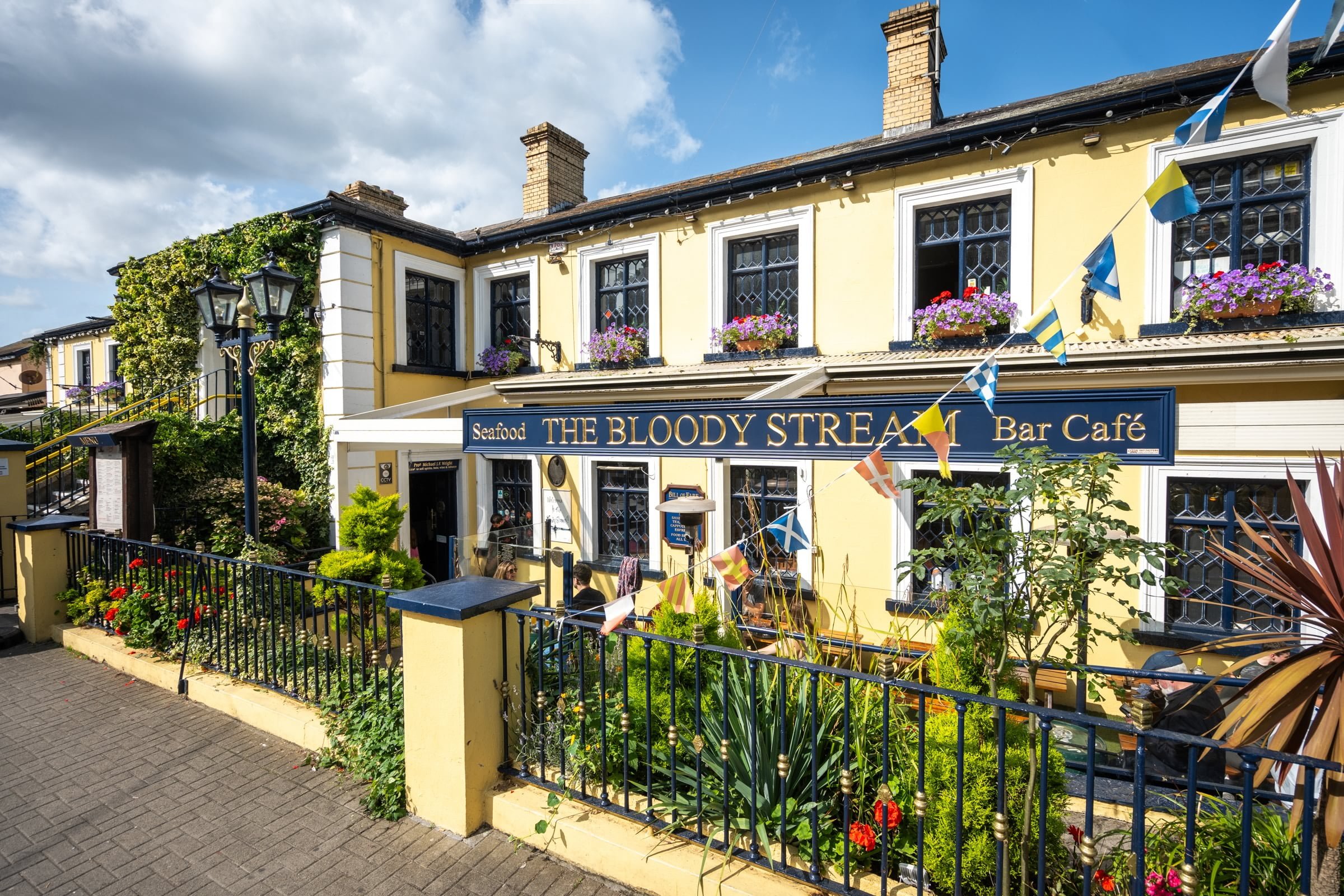 Exterior of a yellow building with a sign reading 'Seafood The Bloody Stream Bar Café,' decorated with multicolored flags, flower boxes on windows, and a black fence with colorful plants and flowers in front, under a partly cloudy sky.