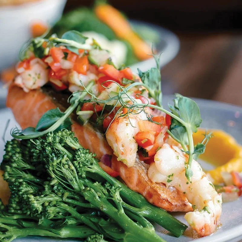 Grilled fish topped with pico de gallo and garnished with microgreens, served with broccoli on a plate.