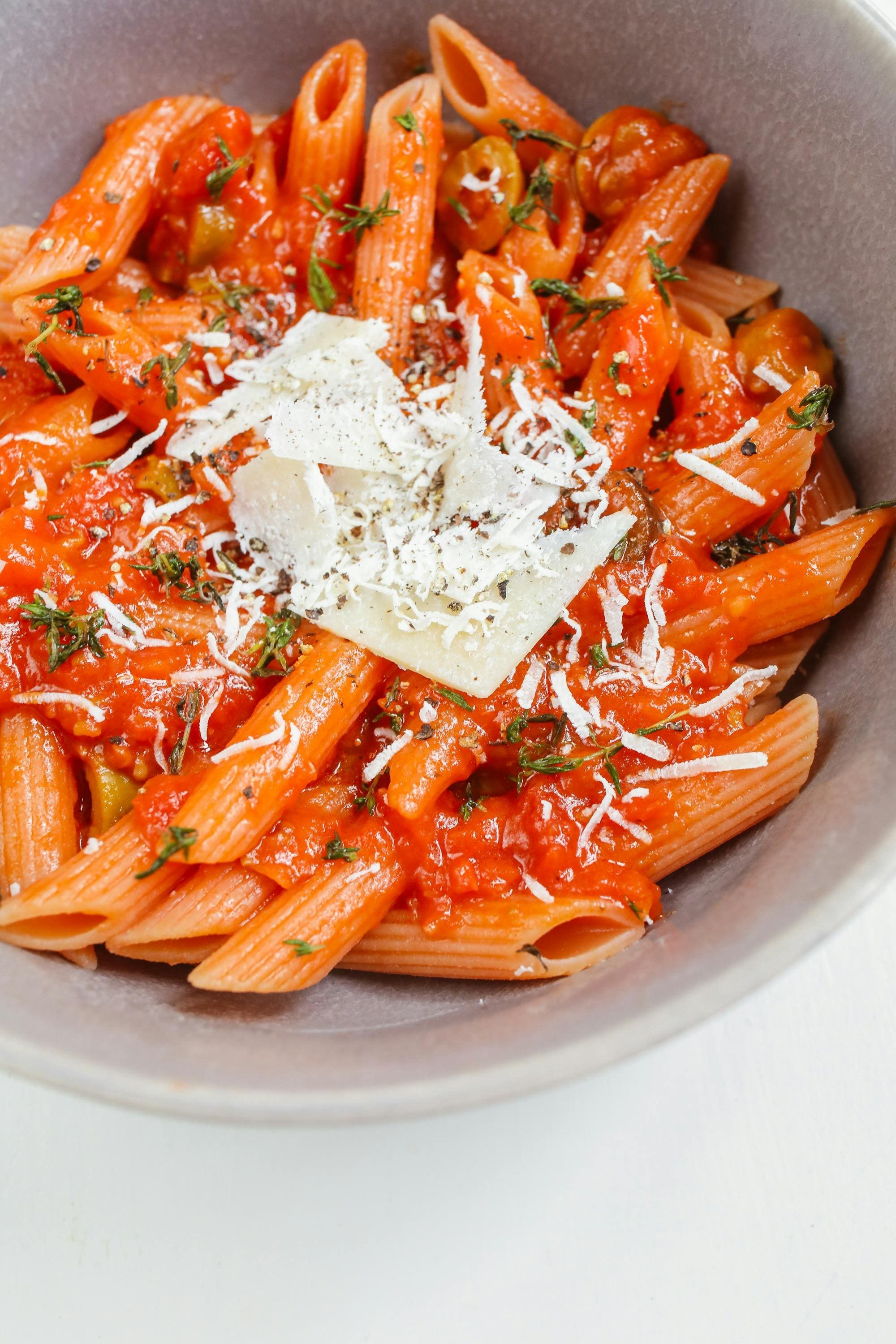 A bowl of penne pasta with tomato sauce, topped with shredded cheese and garnished with herbs.