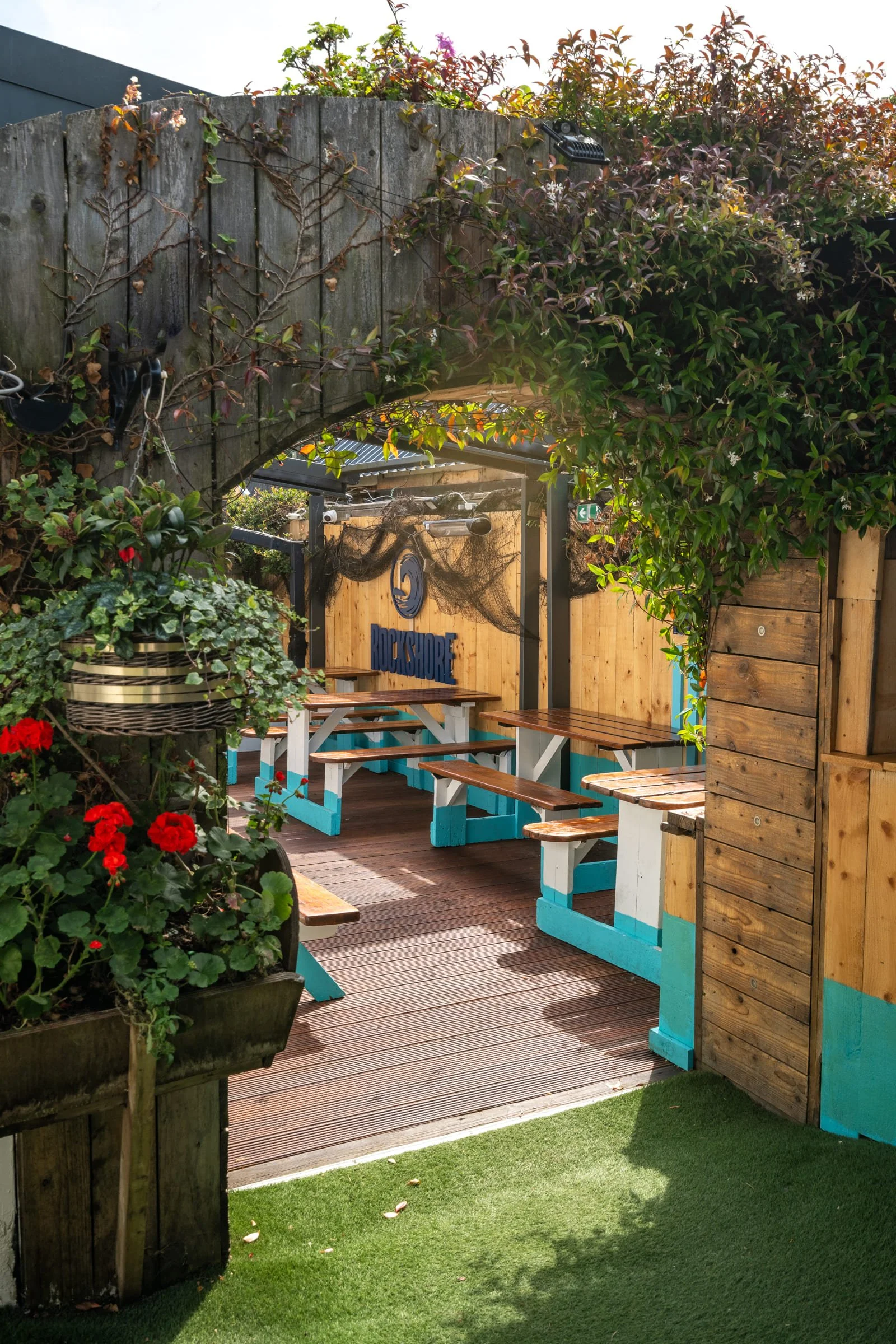 Outdoor seating area with wooden picnic tables and benches, surrounded by greenery, flowers, and wooden fences