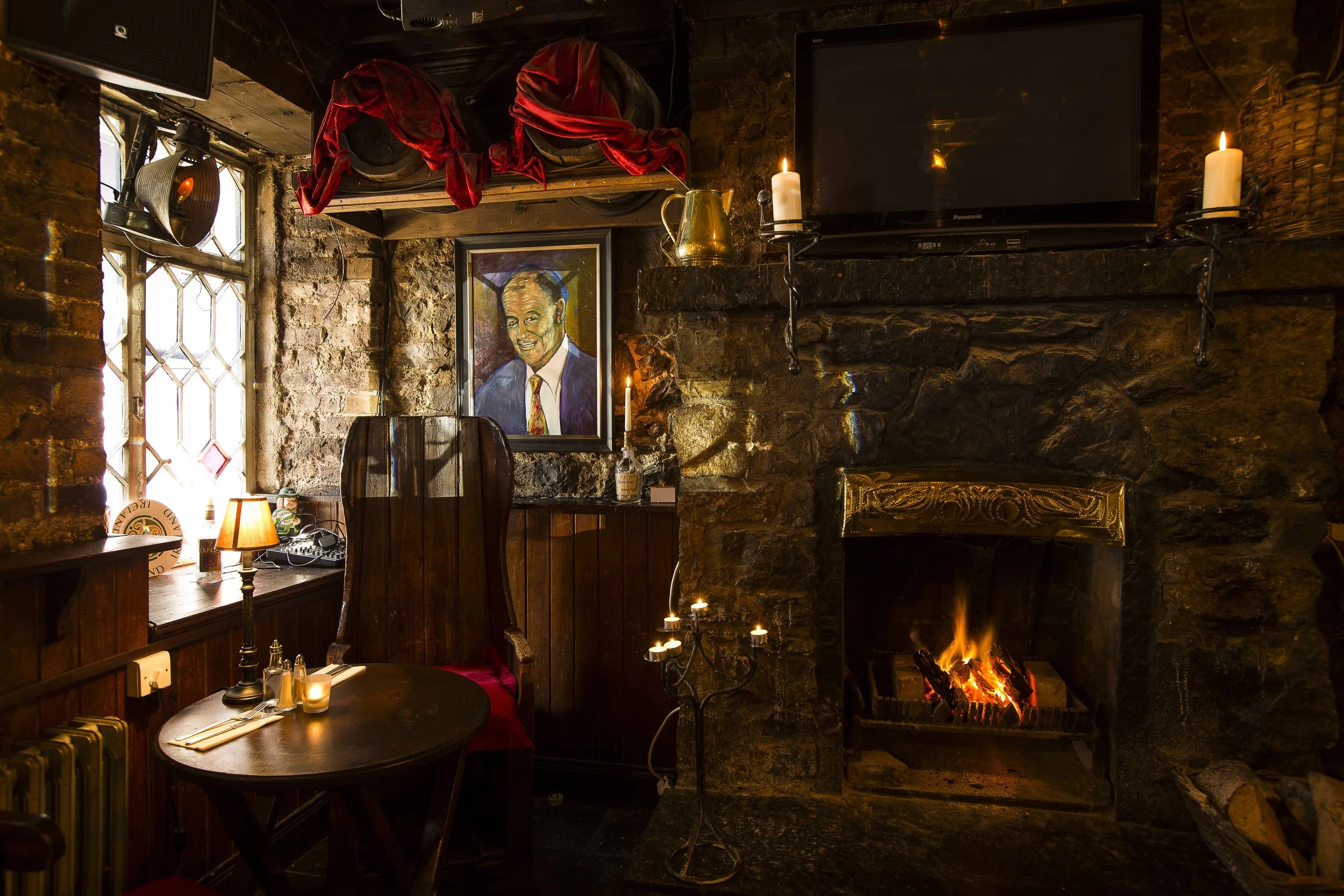 Cozy interior of a rustic pub with a stone fireplace, lit candles, a portrait on the wall, a small table with a lamp and place setting, and a television mounted above the fireplace.