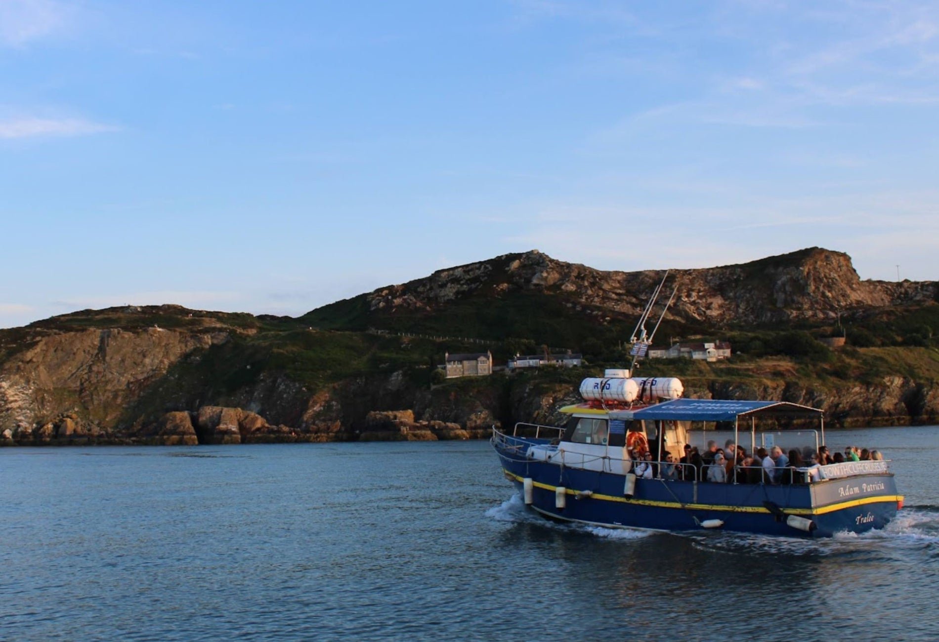 A blue passenger boat with an open upper deck sailing on calm water near a rocky coastline with green hills and houses under a partly cloudy sky.