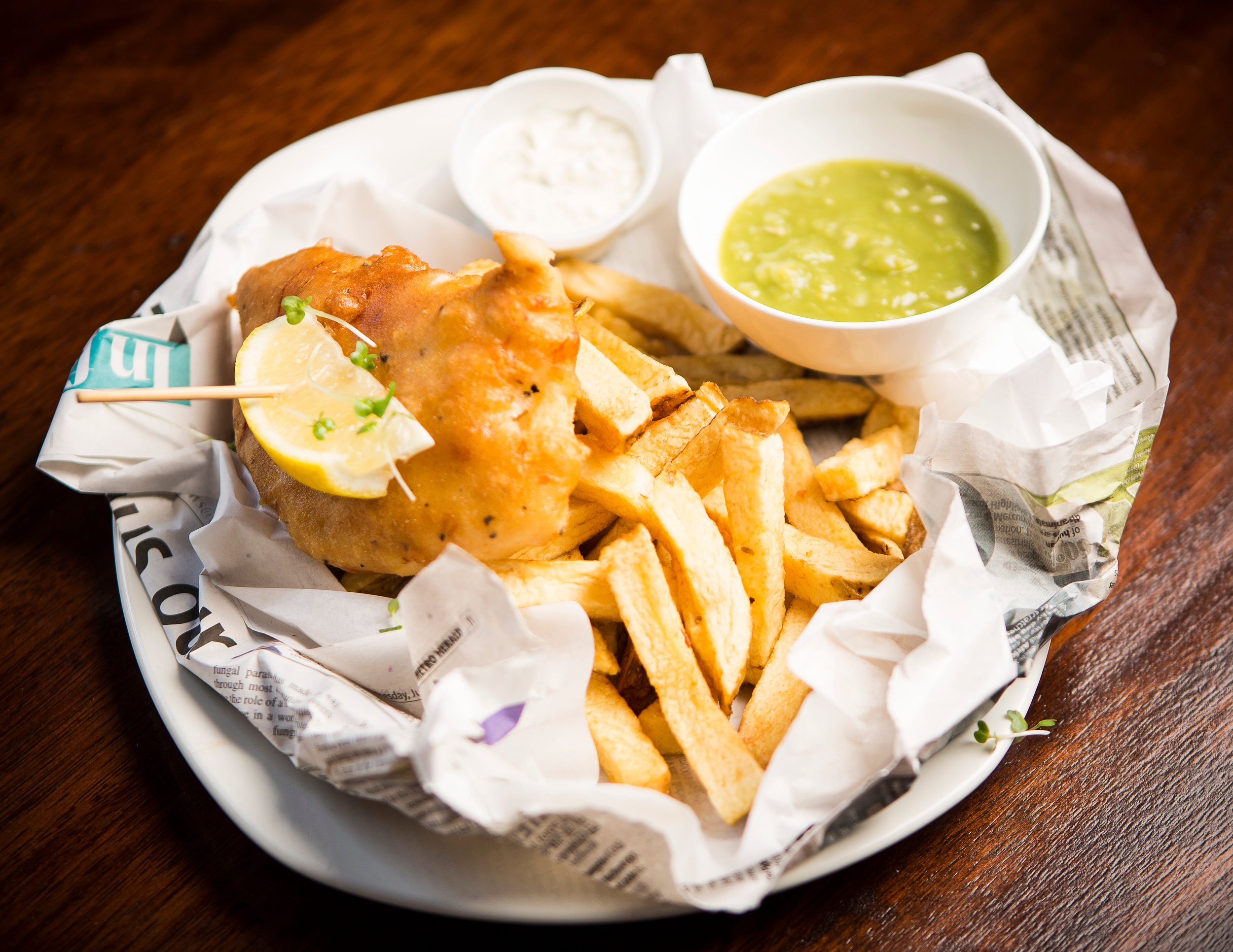 Fried chicken leg with lemon wedge and microgreens, served with French fries, green gravy, and white dipping sauce on a paper-lined plate.