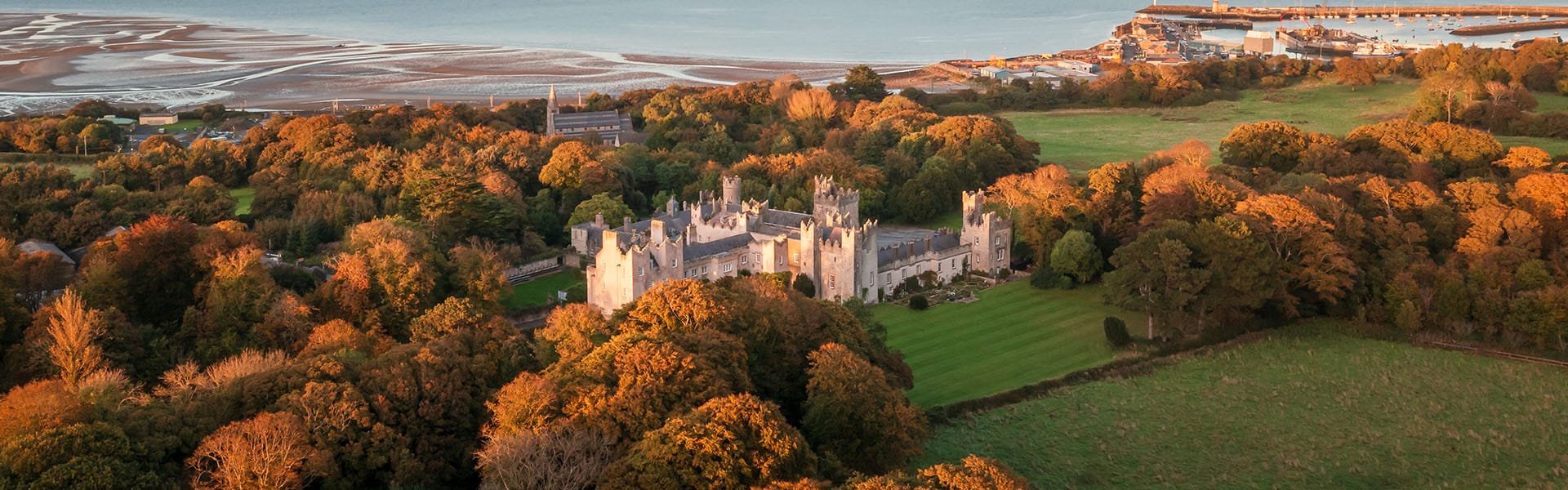 Aerial view of a large castle surrounded by trees with autumn foliage, green fields, and a distant coastline with shoreline and buildings.