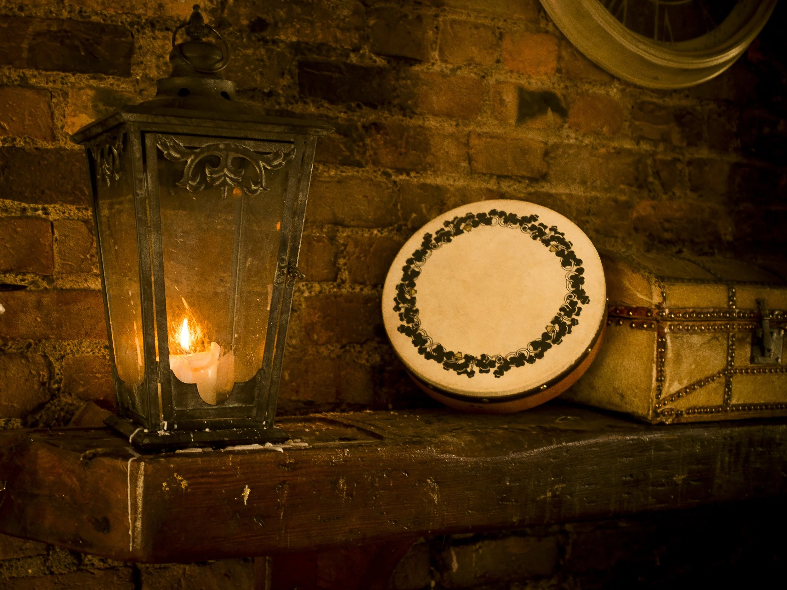 A lit candle inside a black metal lantern on a wooden mantle, with a white decorative drum and a patterned box against a brick wall.