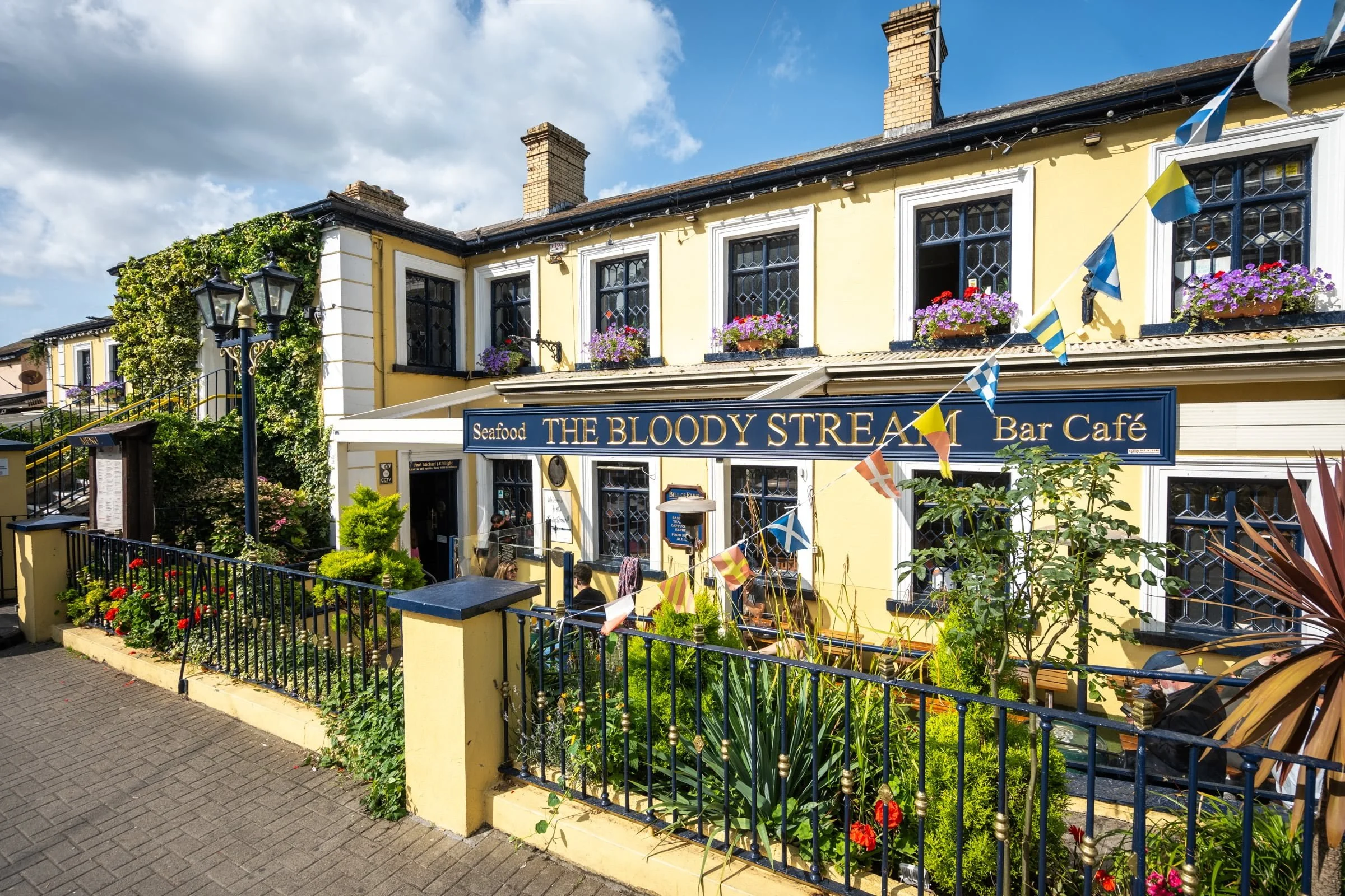 Yellow building with black framed windows, flower boxes, and a sign for The Bloody Stream seafood bar cafe, outdoor seating with people, decorative flags, lush plants, and a blue sky with clouds.