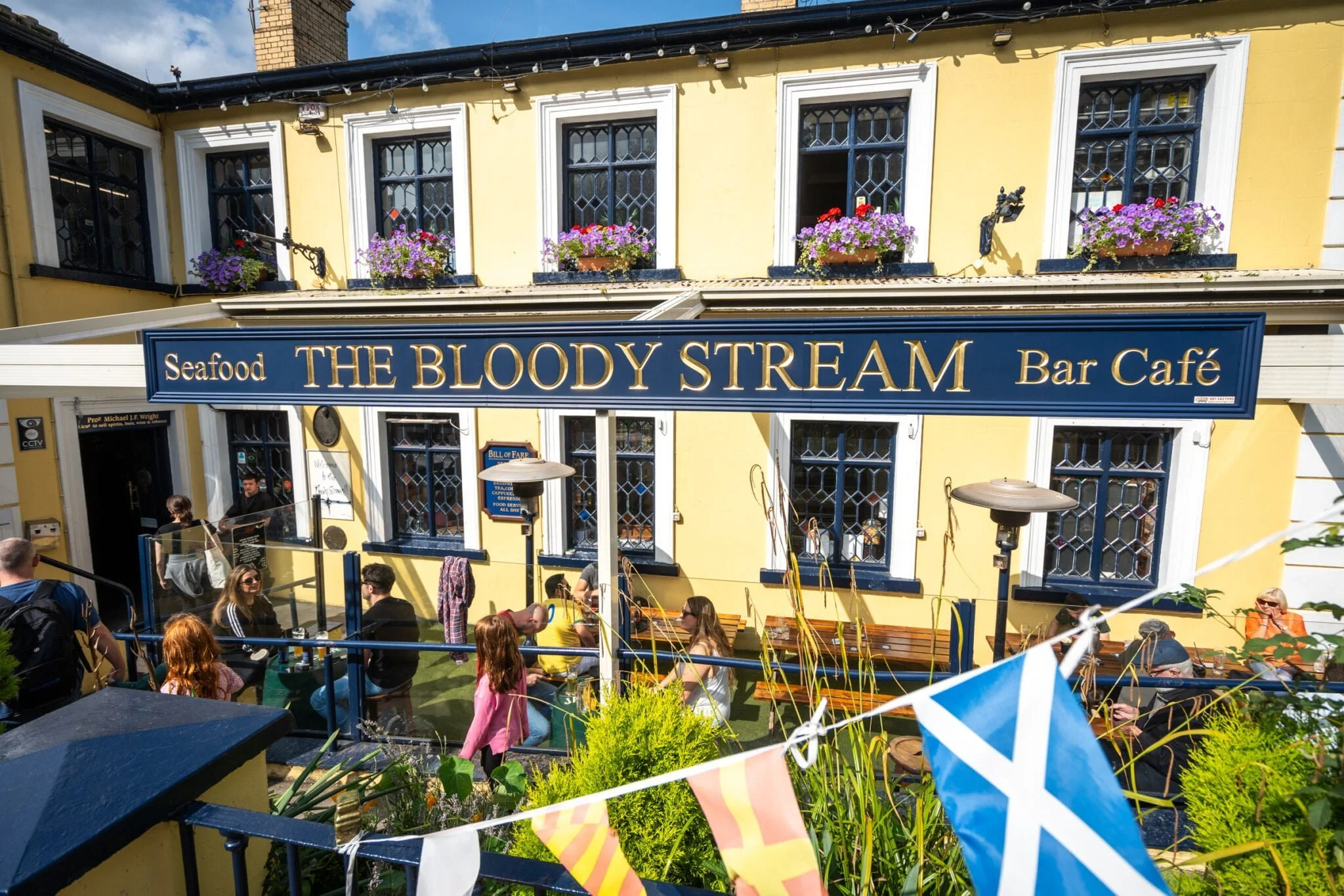 Exterior view of a yellow building with blue and gold sign that says "Seafood The Bloody Stream Bar Café," with patrons sitting outside on benches and tables, some wearing sunglasses, and people entering through a door. Flower boxes with pink and purple flowers decorate the windows.