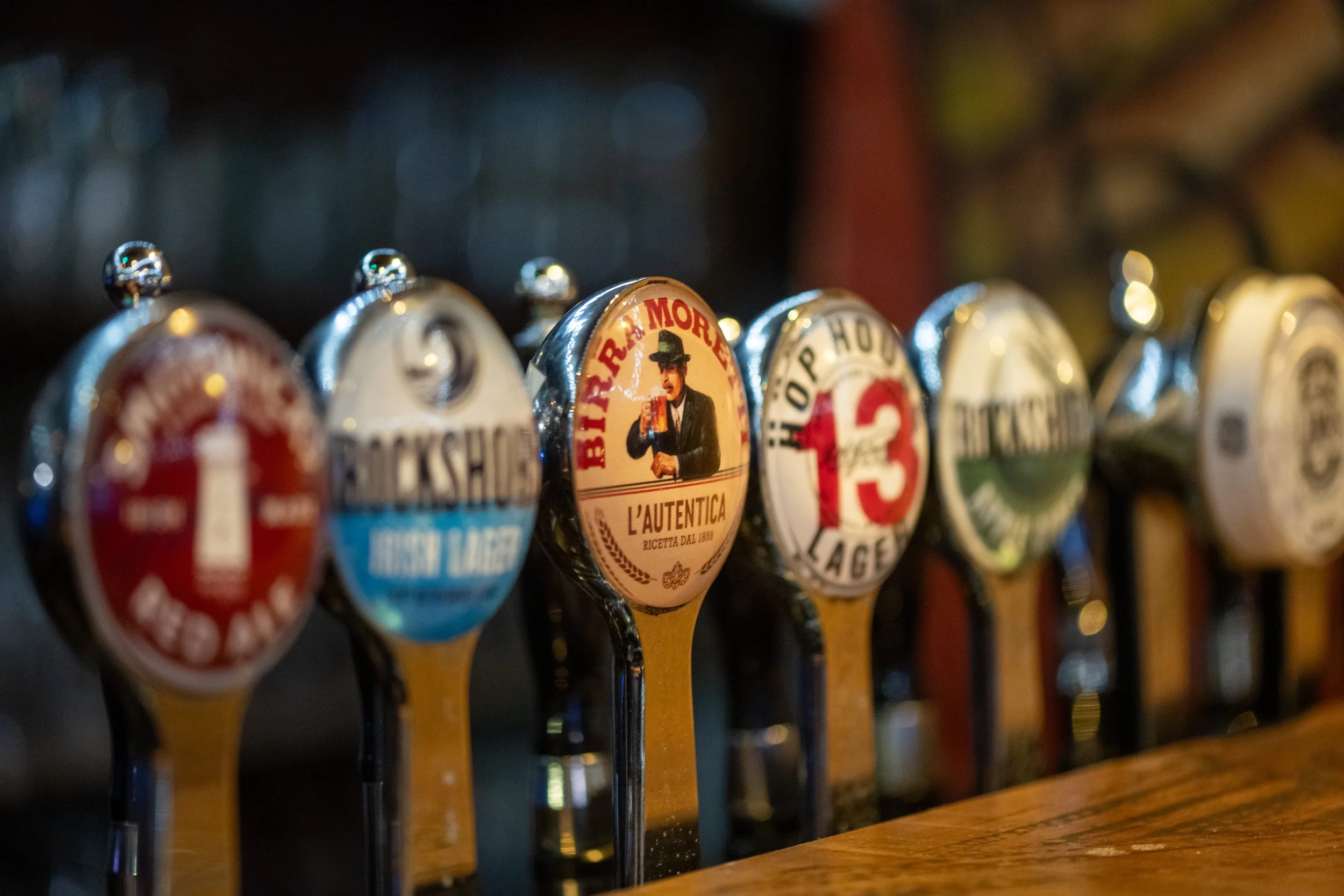 Close-up of beer tap handles at a bar, with various labels including Birra Moretti, Rockshore, and others, in a dimly lit environment.