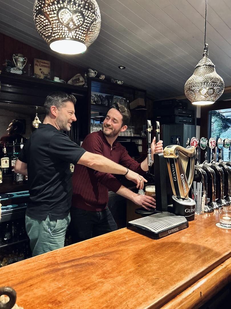 Two men are smiling and chatting behind a bar counter, one of them is pouring a Guinness beer from tap. The bar has decorative hanging lamps and shelves stocked with bottles.