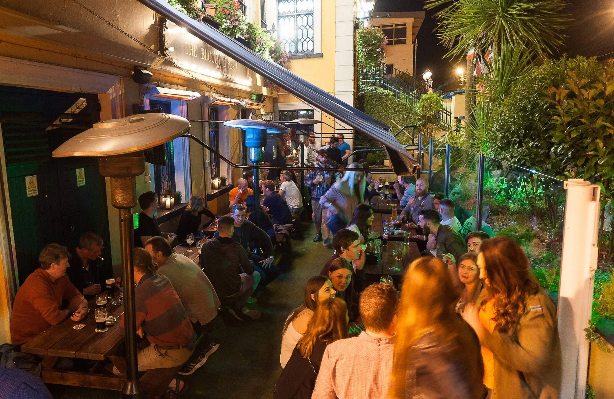 Outdoor patio at a restaurant with groups of people sitting at tables, some standing and talking, illuminated by warm lighting, with plants and greenery surrounding the area at night.