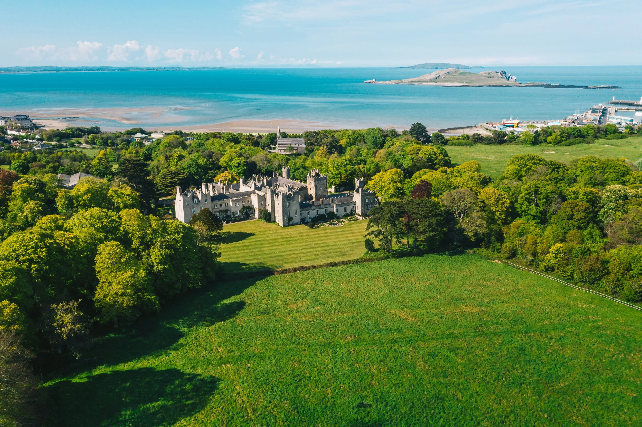 Aerial view of a historic castle surrounded by lush green trees and open grassy fields, with a coastline and the ocean in the background.