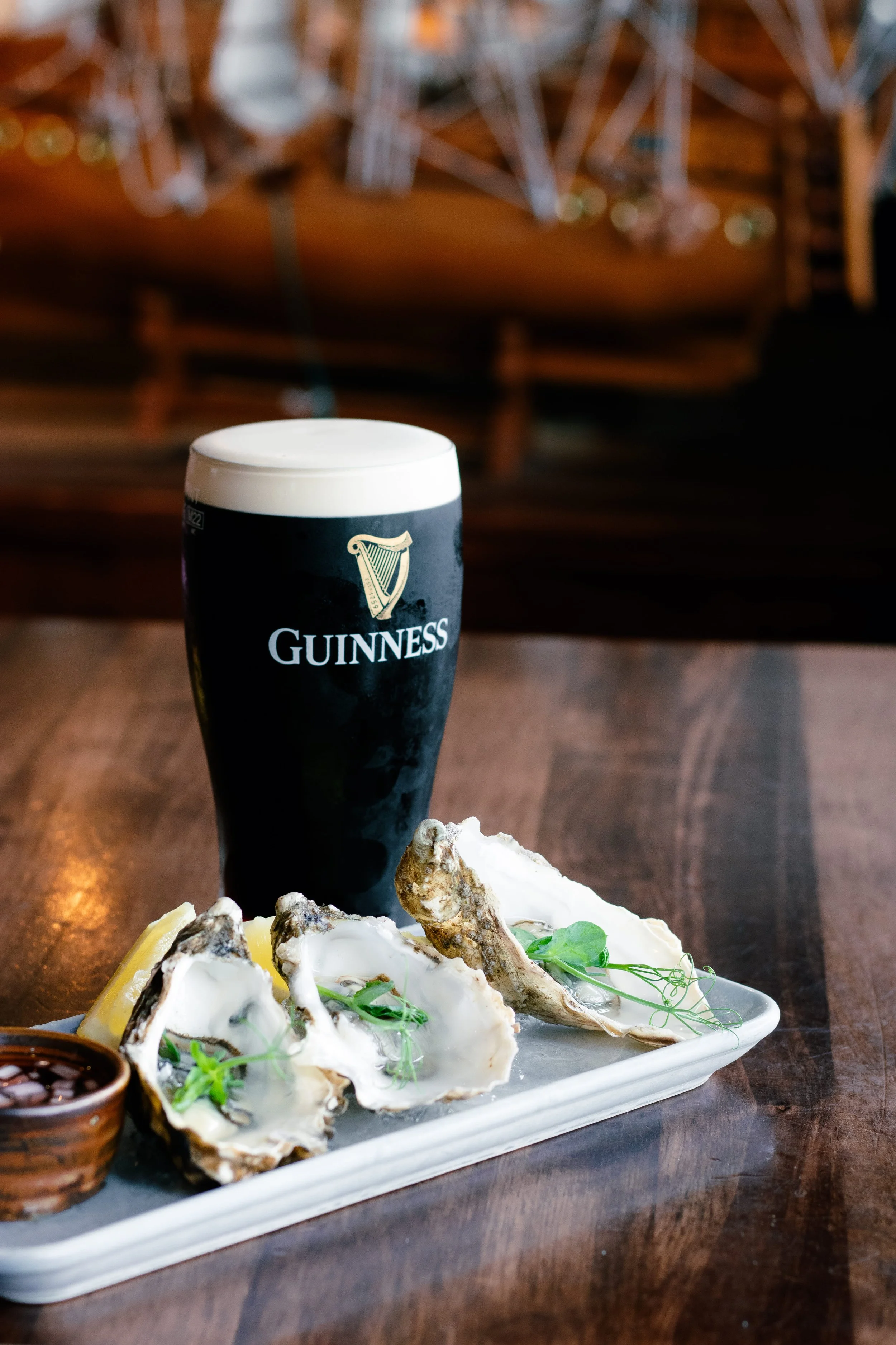 A pint of Guinness stout beer with a creamy head and a plate of raw oysters garnished with green herbs and lemon wedges, with a small bowl of dipping sauce on a wooden table.