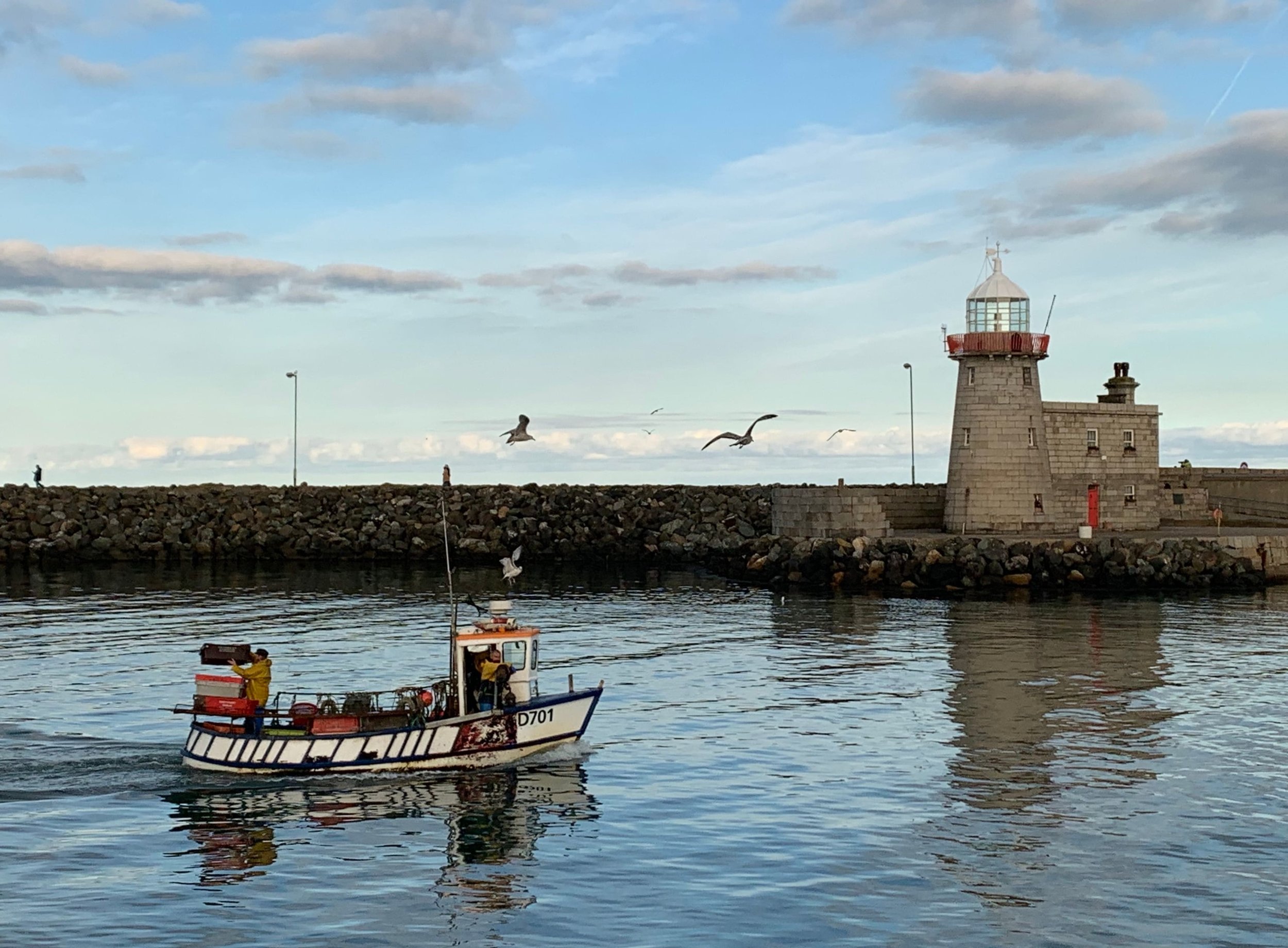 A lighthouse on a stone pier with a boat sailing nearby, seagulls flying in the sky, and a person walking in the distance on the pier, under a partly cloudy sky.