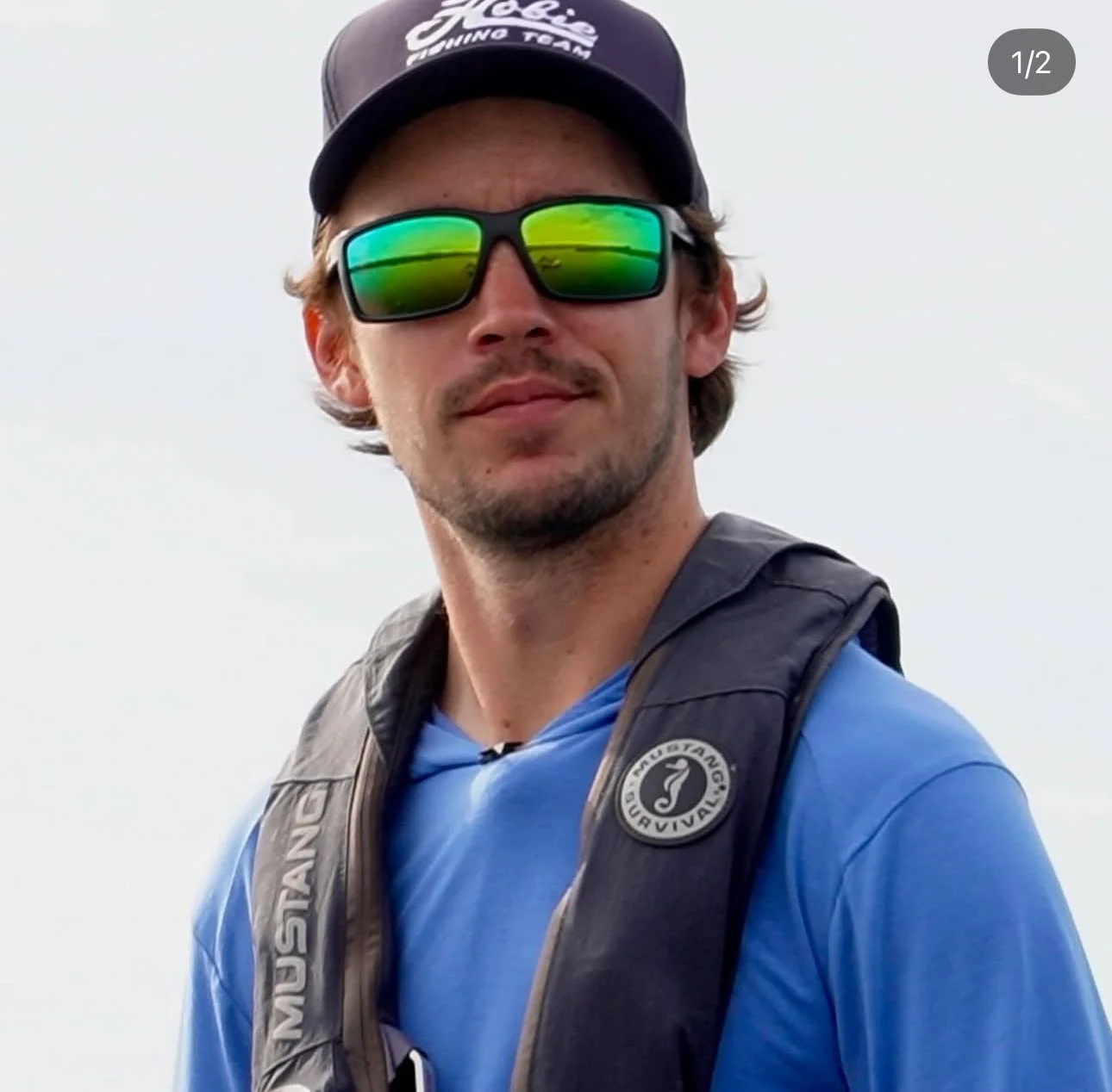 A man wearing a hat, sunglasses with green reflective lenses, and a blue shirt, carrying a black backpack with a circular logo.