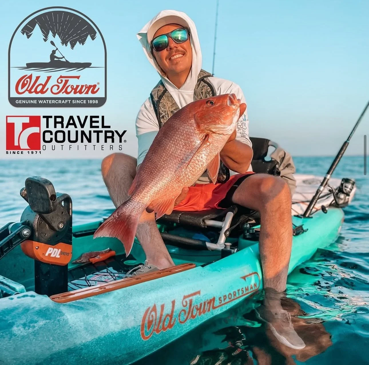 A man in fishing gear sitting in a kayak on the water, holding a large red fish, with fishing rods and equipment visible on the kayak. The kayak has the logo 'Old Town Sportsman' and the image includes logos for Old Town and Travel Country Outfitters