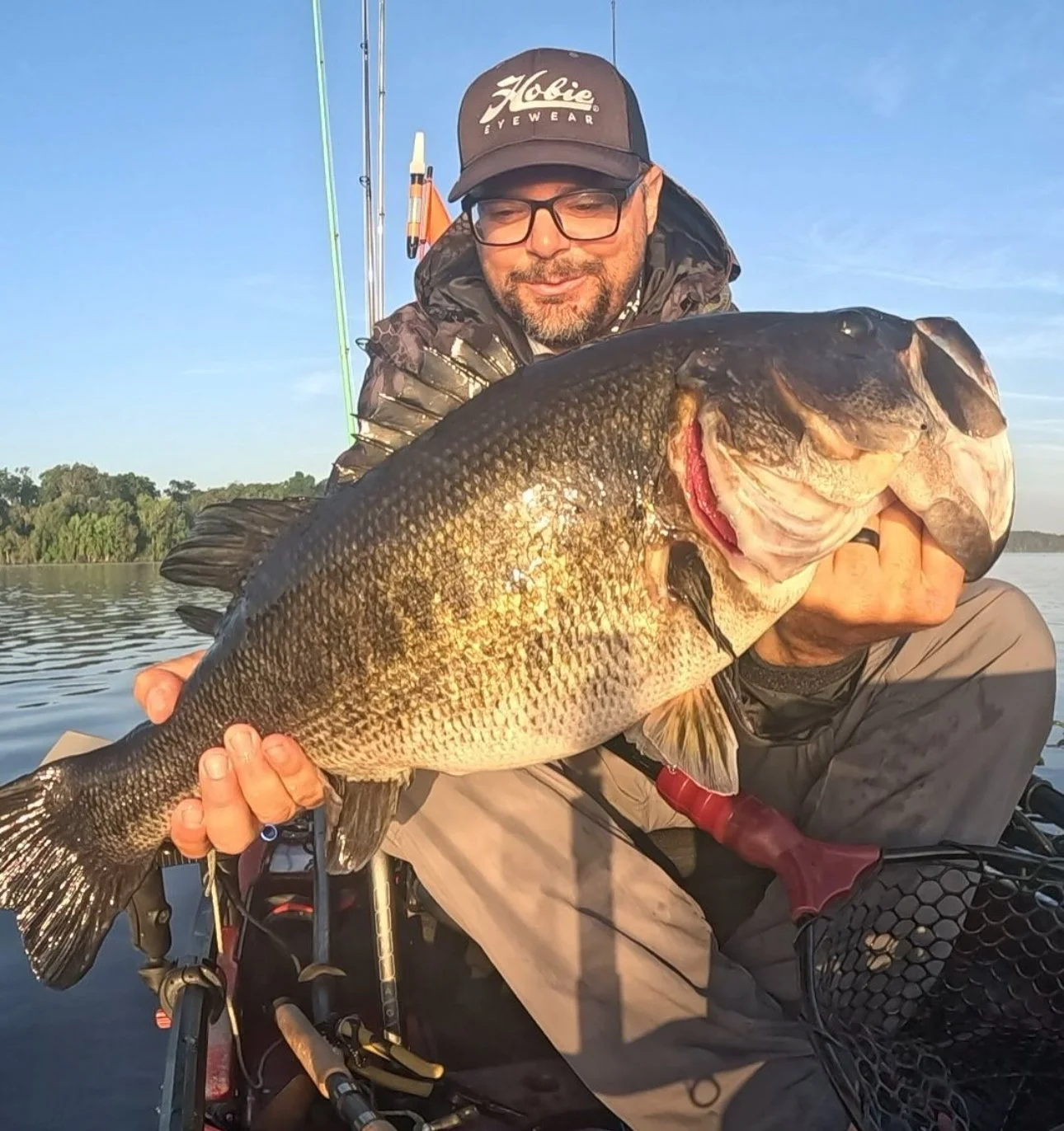 Man in a hat and glasses holding a large fish on a boat in a lake during sunset.