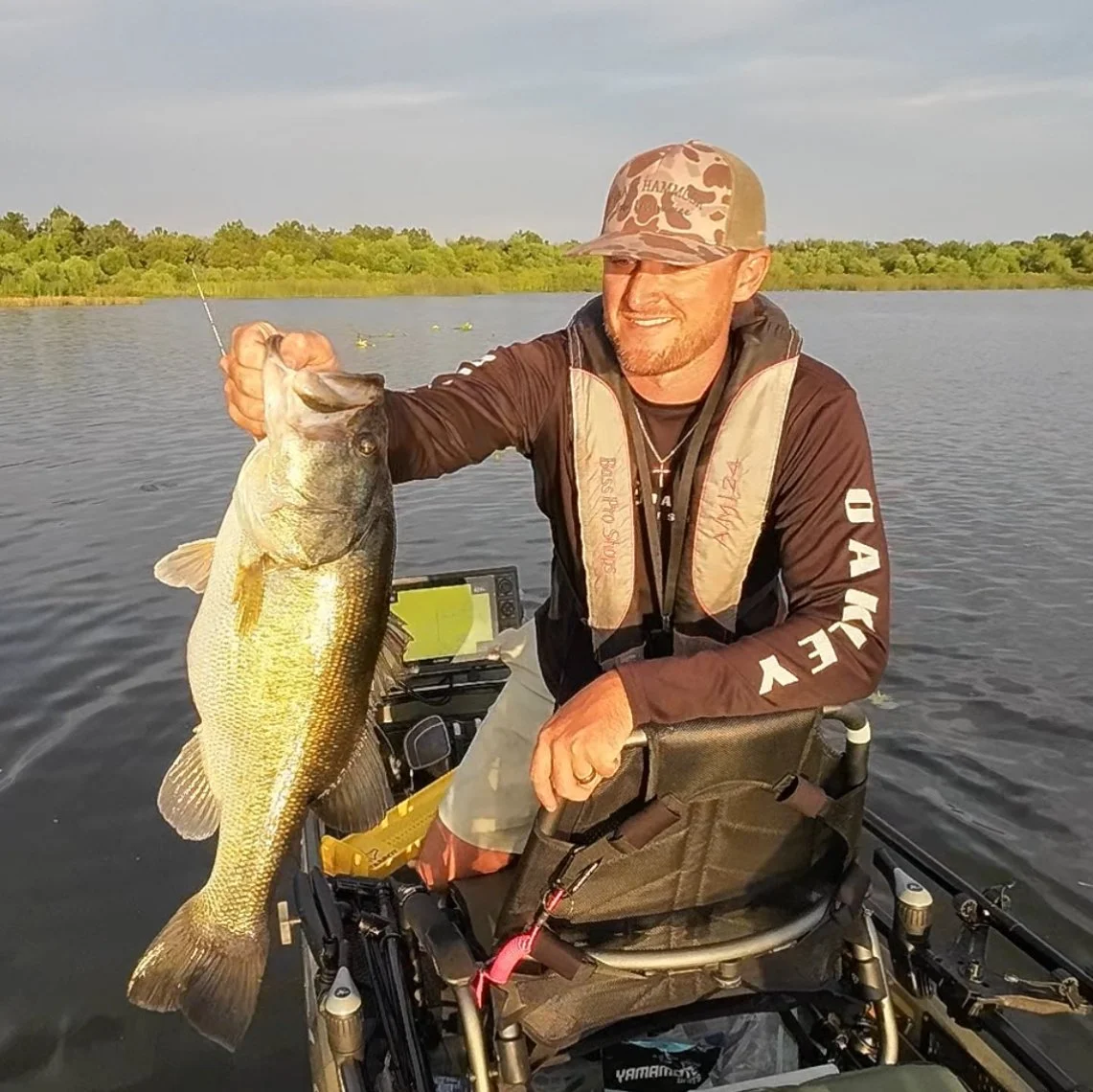 A man in a wheelchair on a boat holding a large fish he caught, with a body of water and green trees in the background.