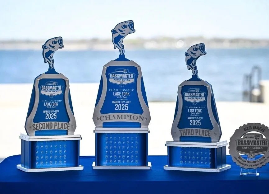 Awards for a kayak bass fishing competition, including first, second, and third place trophies, with a backdrop of a body of water. Kayak Bass Fishing in Florida