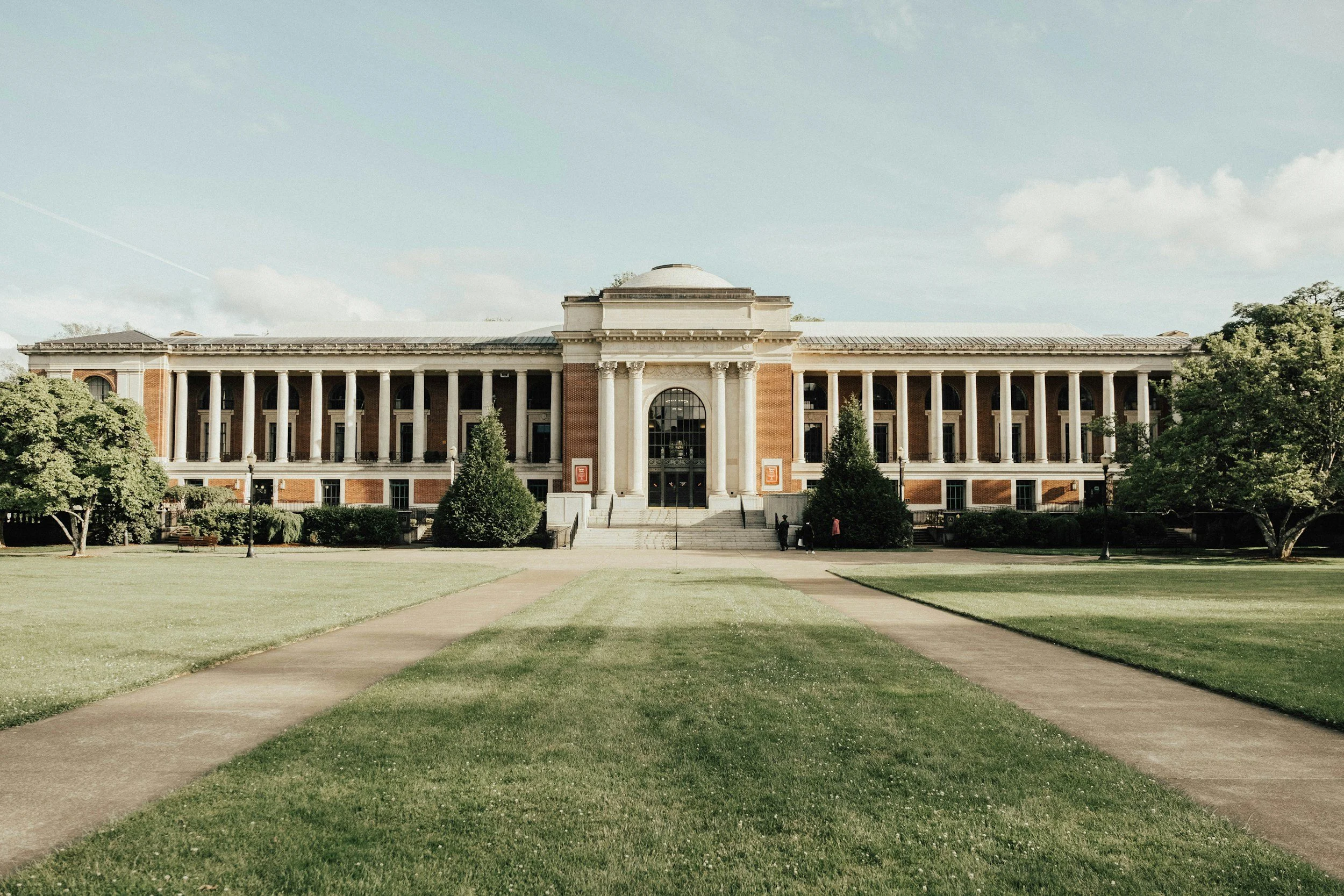 Large historic academic building with columns, brick walls, and a rounded central section, surrounded by a well-maintained lawn and trees.