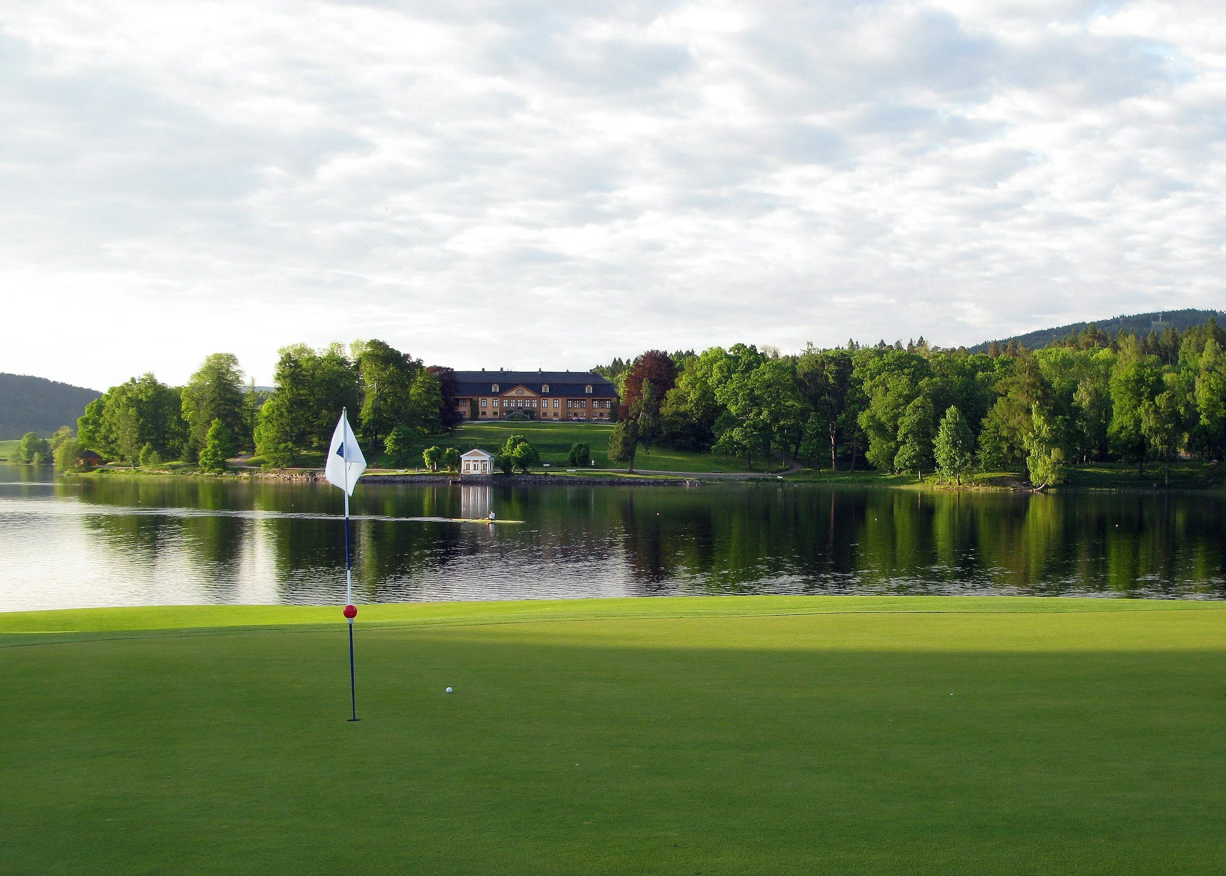 A golf course with a putting green, flag, and golf ball, overlooking a lake with a large building surrounded by trees in the background under a cloudy sky.