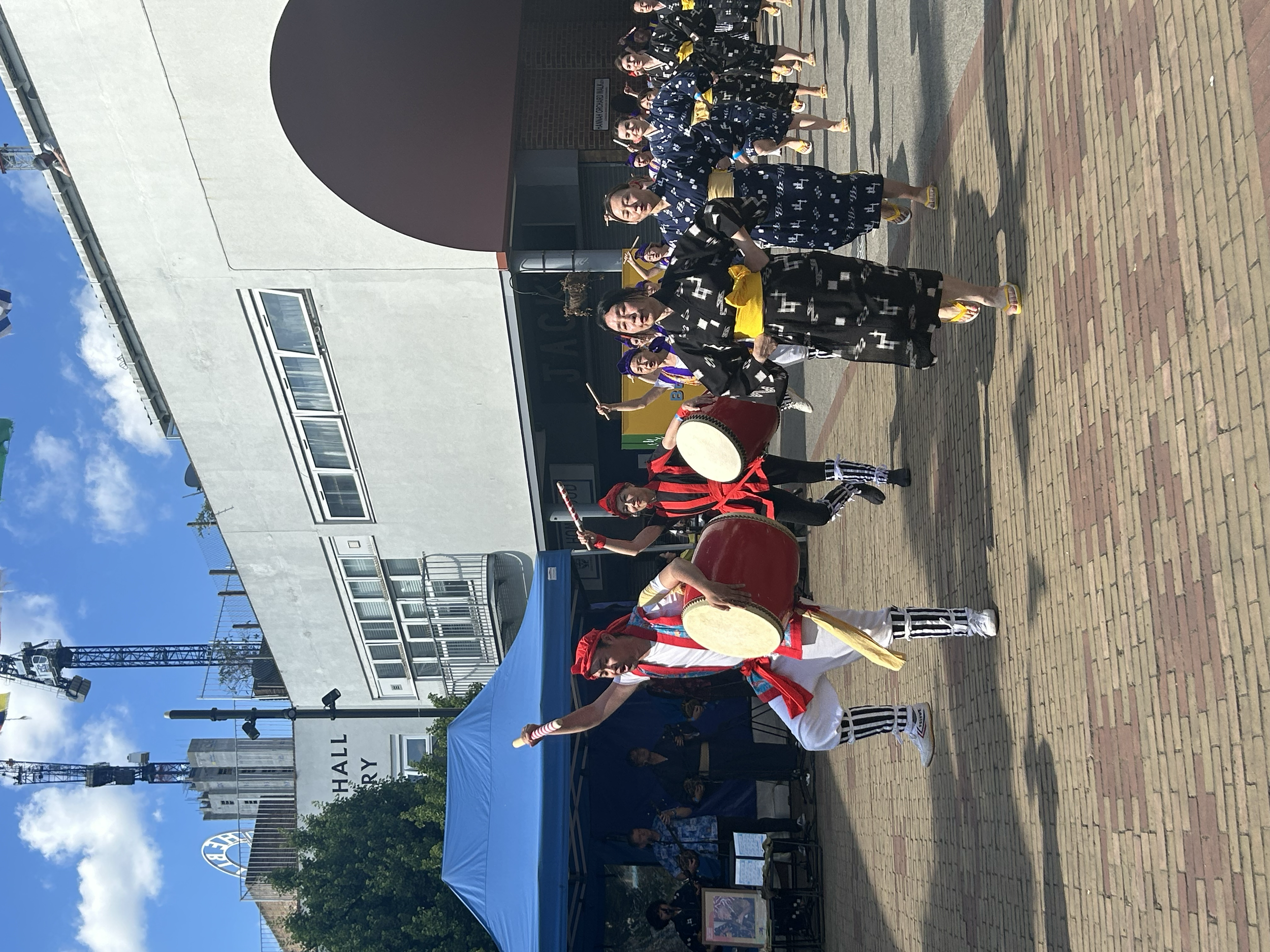 Group of musicians playing traditional Japanese taiko drums outdoors during a festival, with some participants wearing black and yellow and others in red and white costumes.
