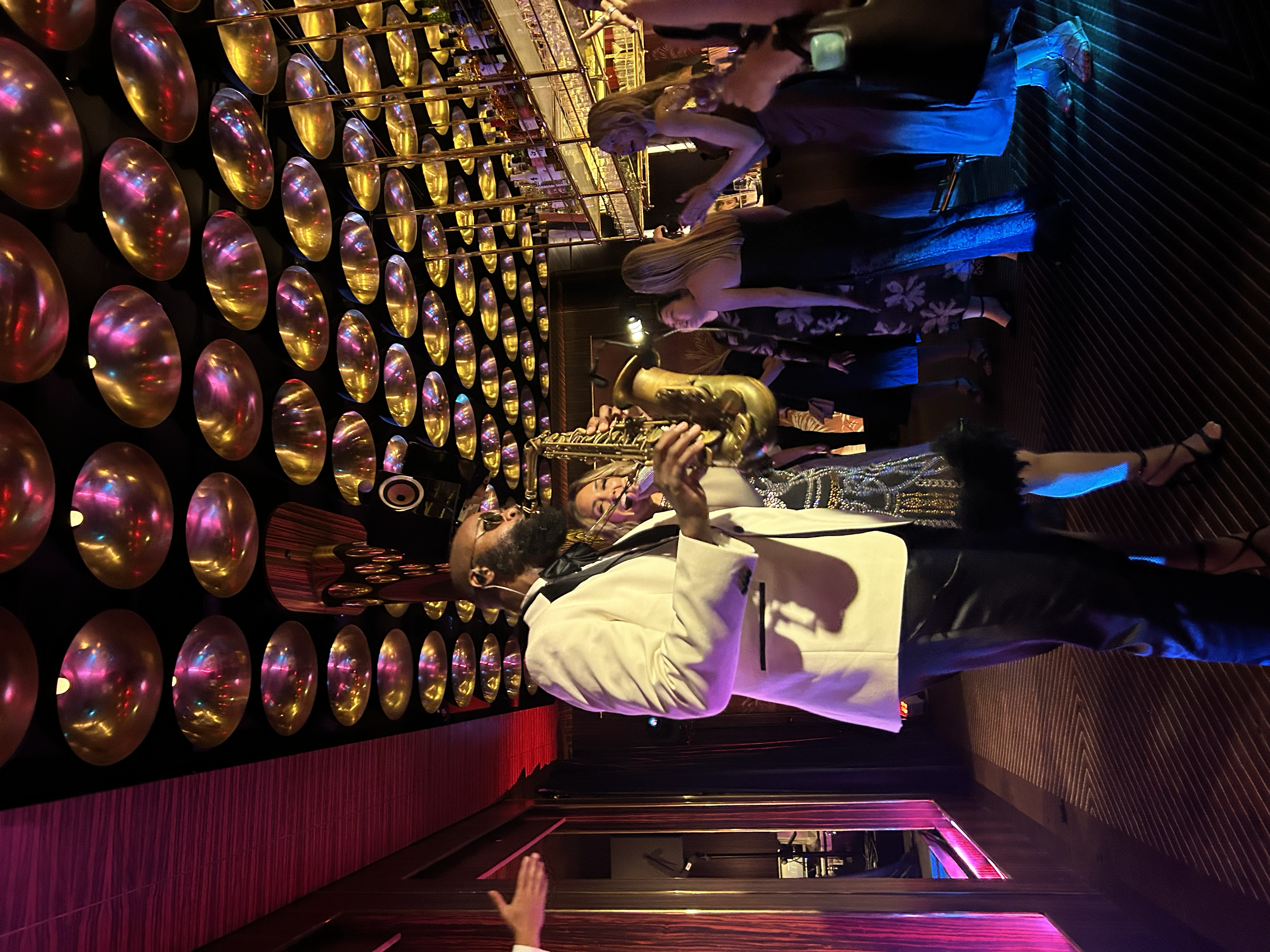 A man in a white tuxedo jacket playing a saxophone at a lively indoor party or event, with other guests in elegant attire dancing and socializing, illuminated by colorful lights and decorated with gold and black circular objects on the wall.