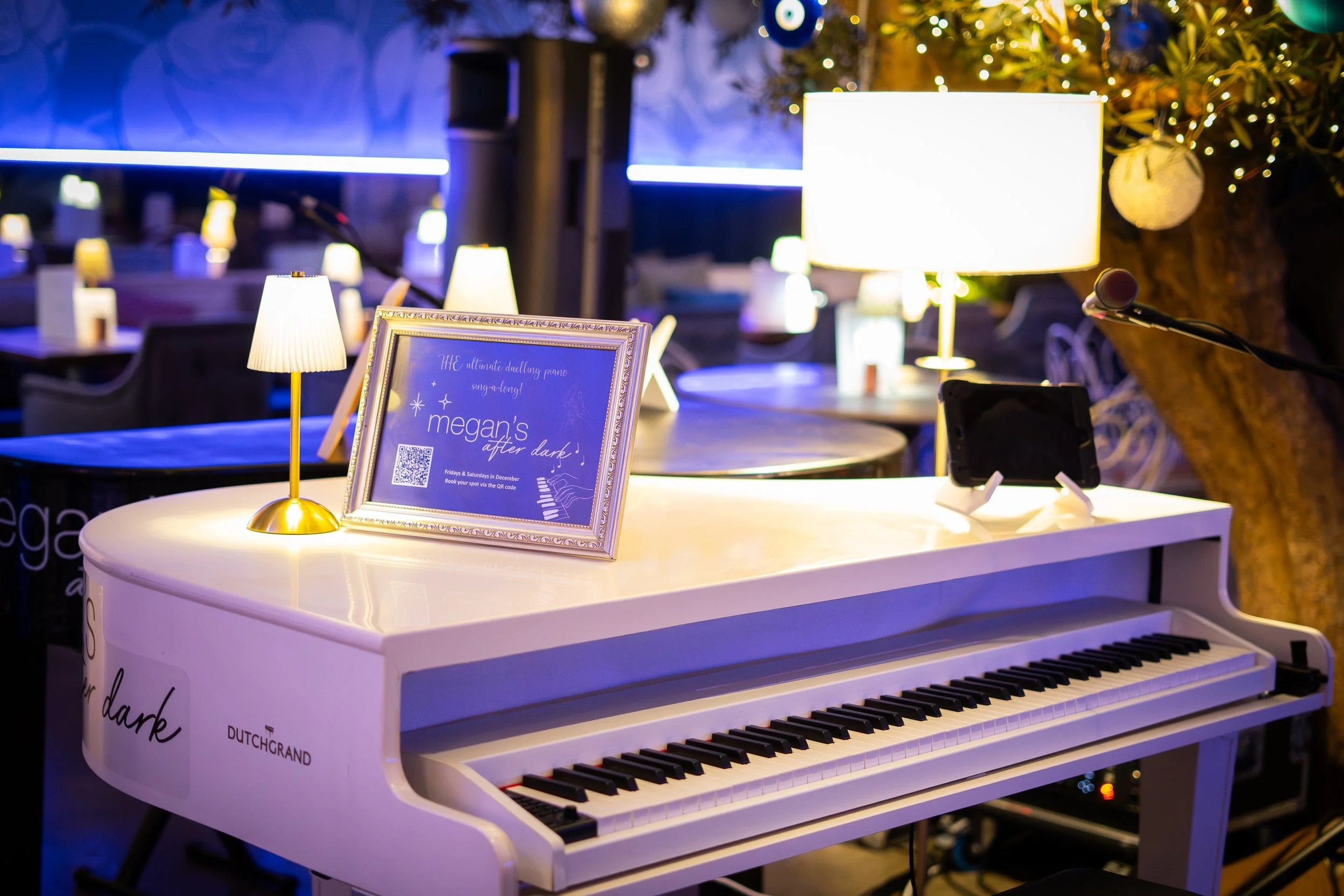 A white piano with a sheet of music or poster on top and a small lamp, in a dimly lit restaurant or event space decorated for the holidays with a Christmas tree and ornaments in the background.