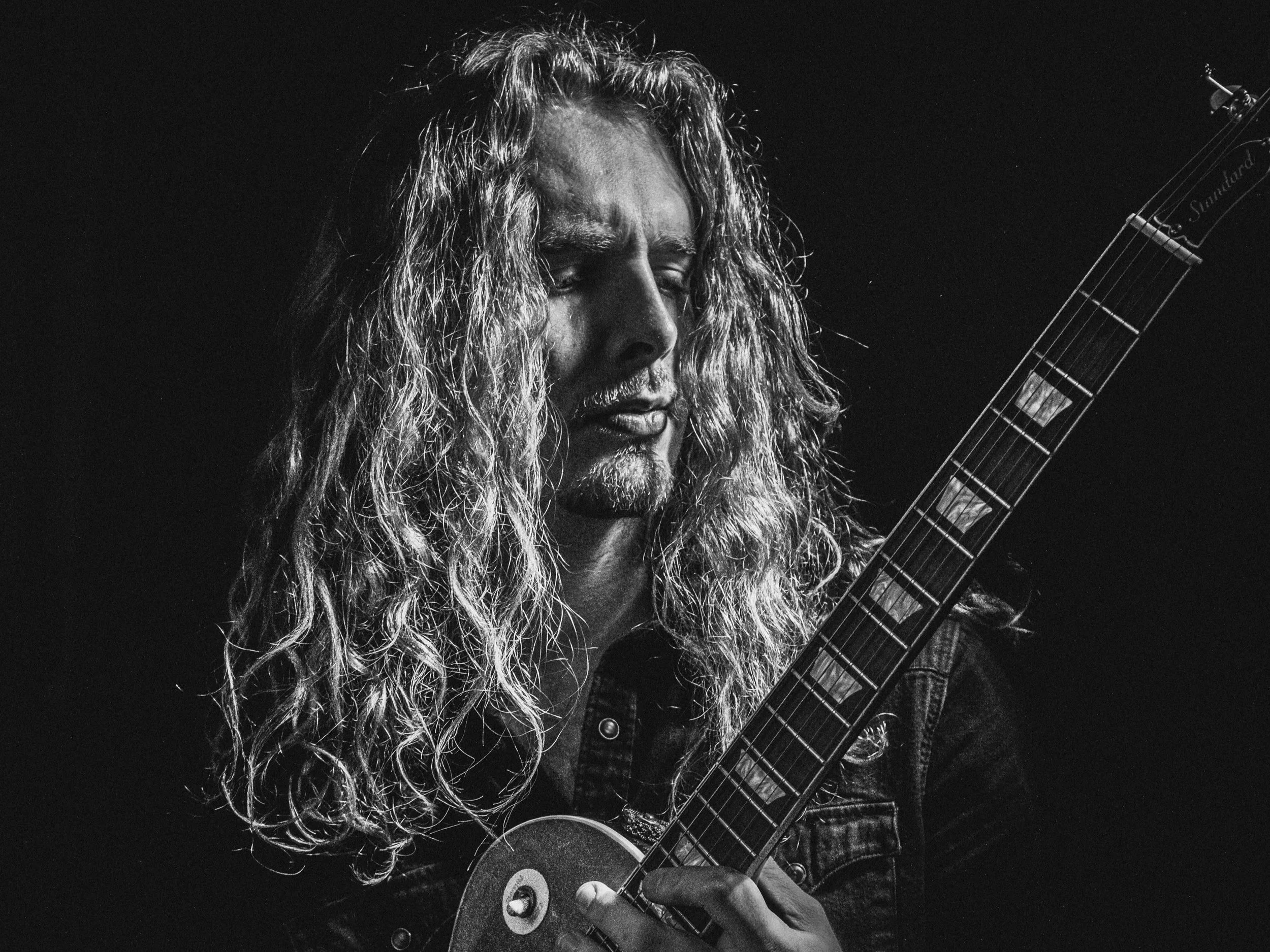 A man with long, curly hair playing an electric guitar, with a focused expression, in black and white.