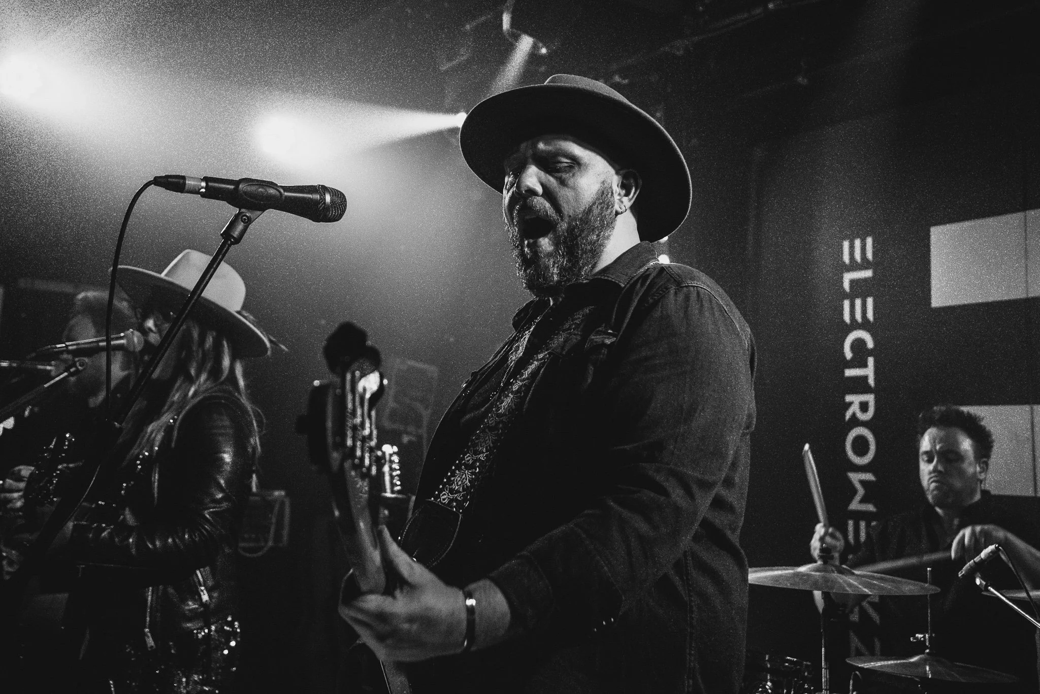 A black and white photo of a male musician singing and playing guitar on stage, wearing a wide-brimmed hat and a dark jacket, with a female singer and a drummer in the background.