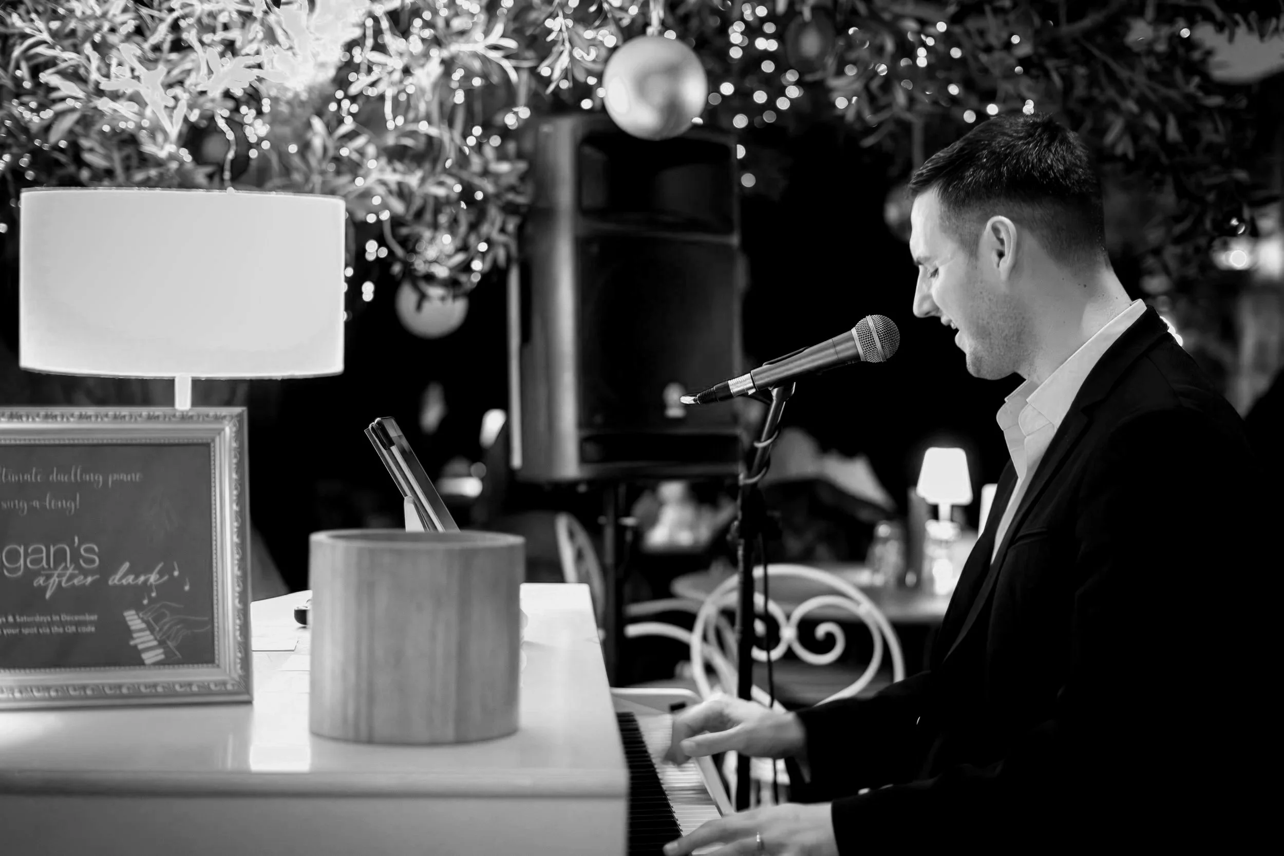 Man playing piano and singing into microphone at a decorated venue with Christmas ornaments and lights.