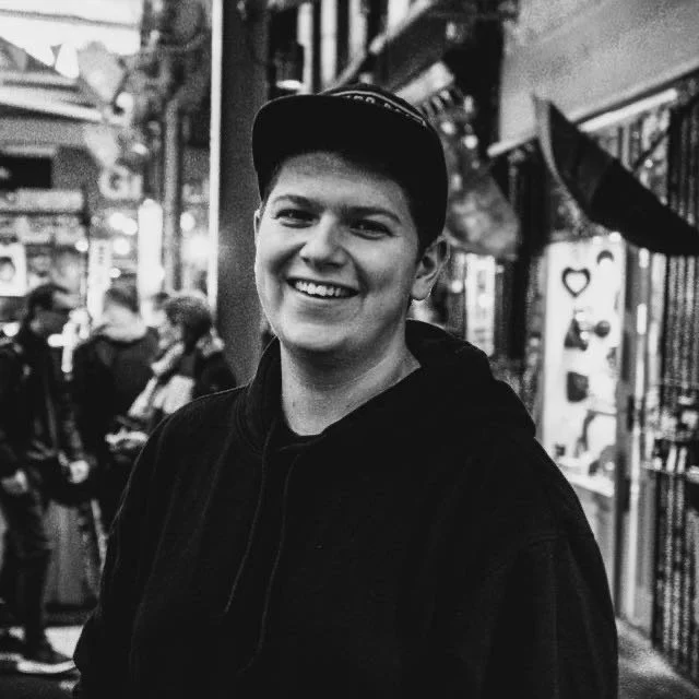 Young man smiling in a busy city street at night, wearing a black hoodie and baseball cap, with shops and people in the background.