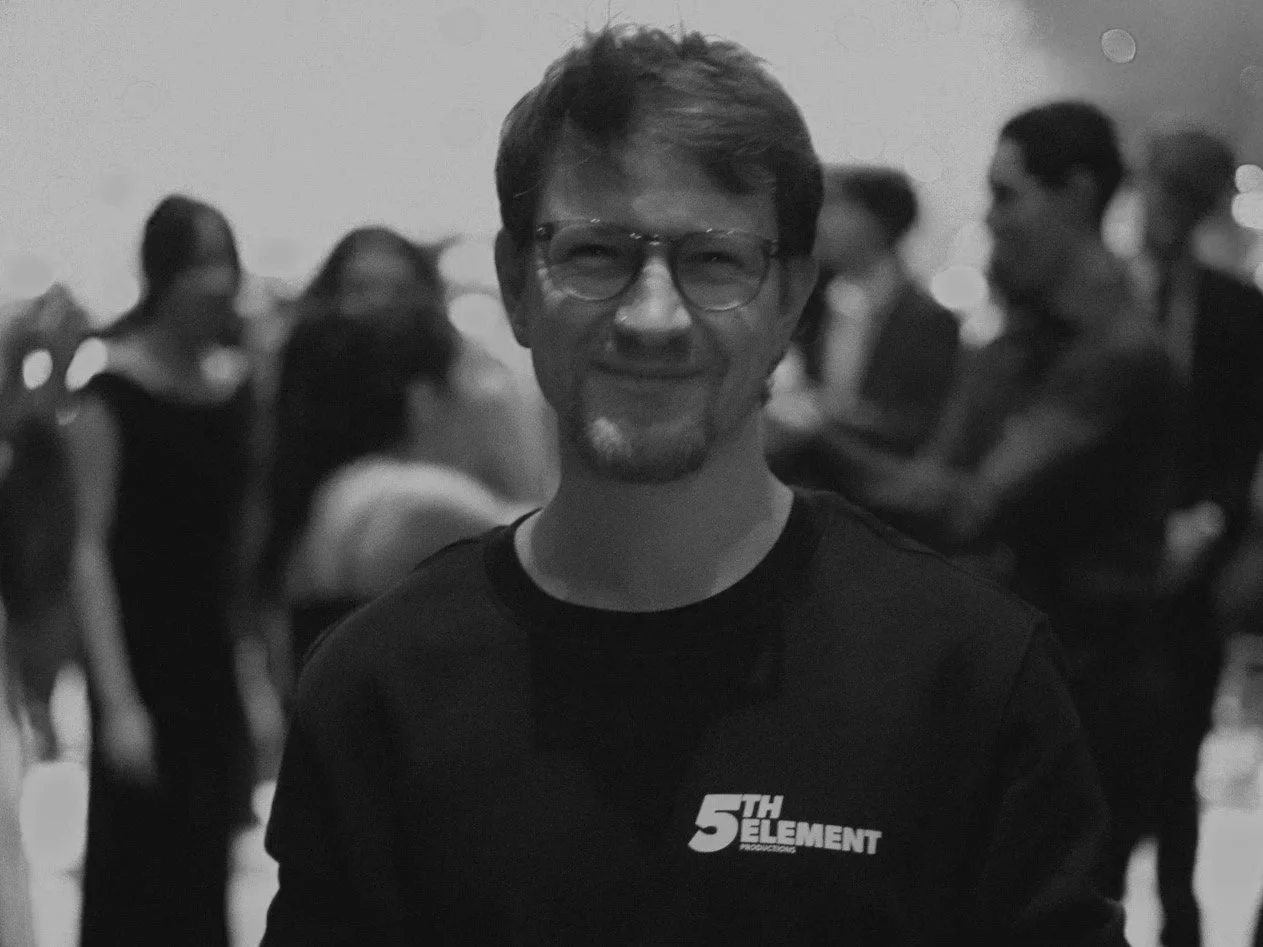 A man with glasses smiling at a social gathering, with several people in the background.