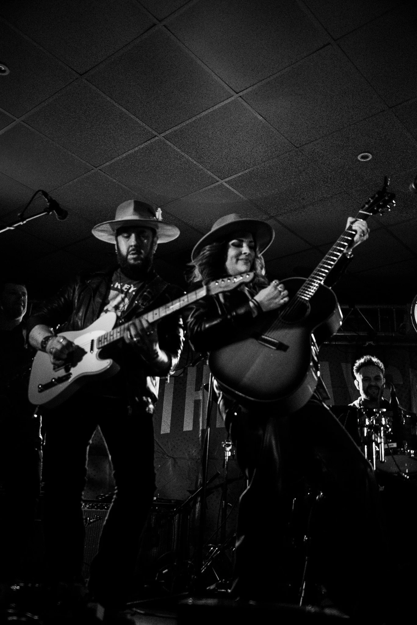 Black and white photo of three musicians on stage, playing guitars and drums, with women in wide-brimmed hats, leather jackets, and a dark ceiling with squares.