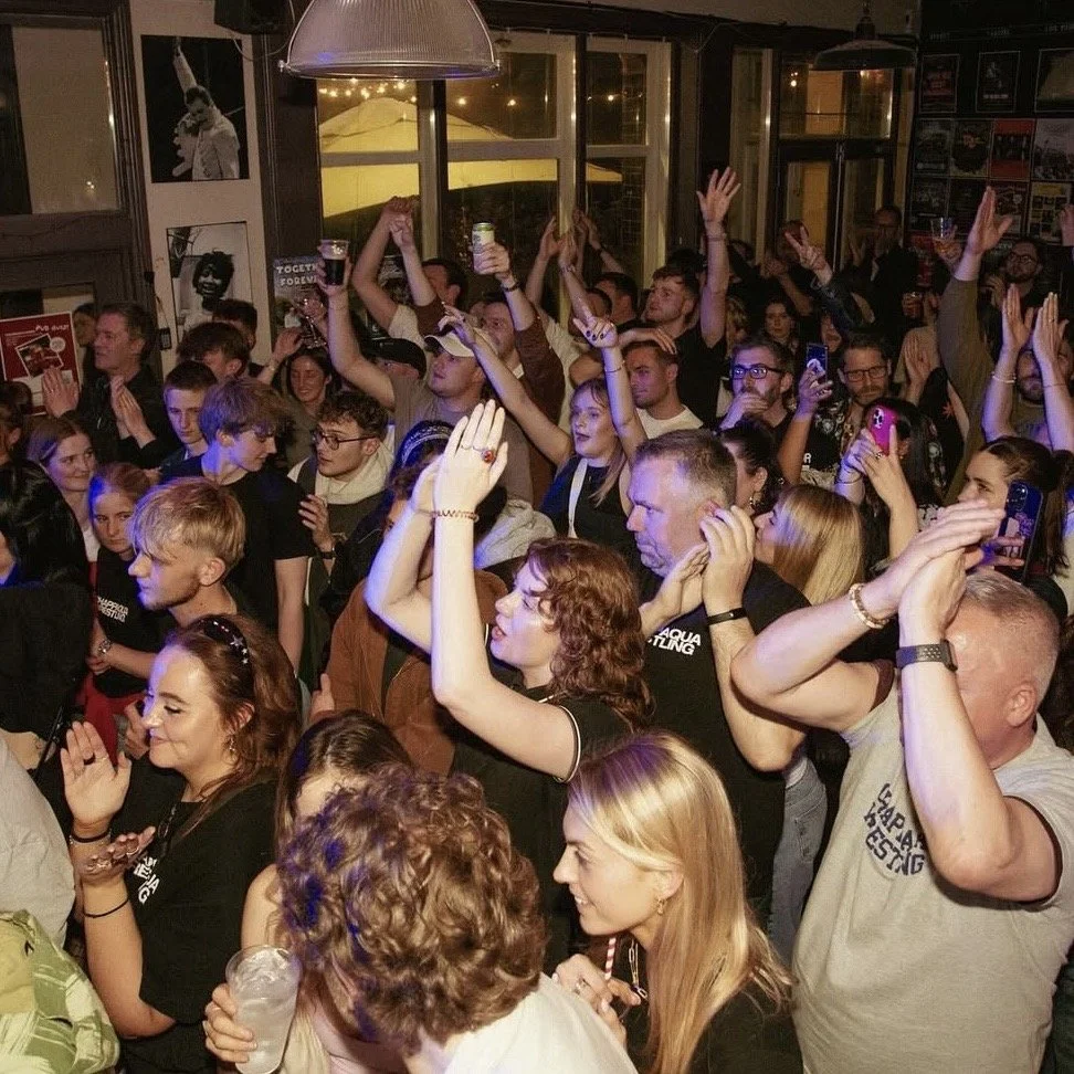 A crowded indoor concert or party scene with many people dancing and enjoying the event, some holding drinks and taking photos.