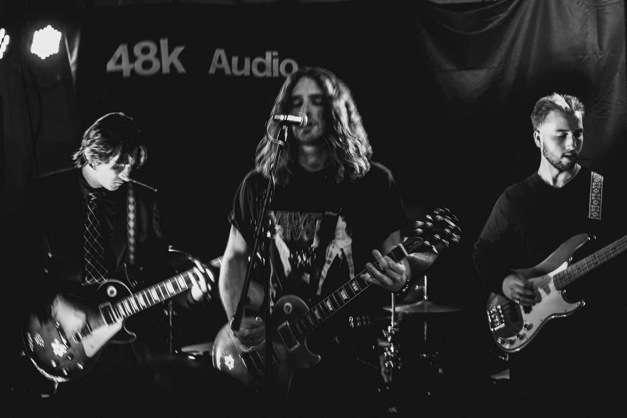 A black and white photo of three musicians performing on stage with guitars, with a sign in the background that reads '48k Audio.' The center musician is singing into a microphone and playing an electric guitar, while the other two are playing guitar