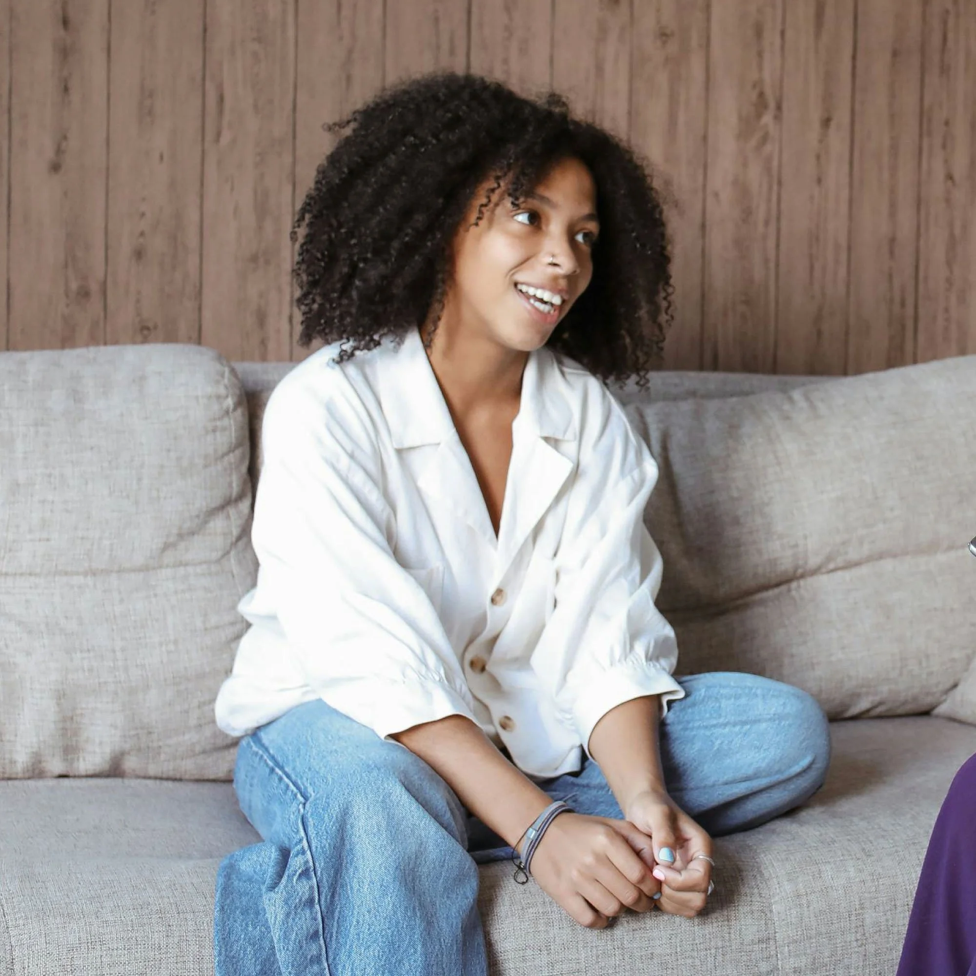 A young woman with curly hair sitting on a beige couch, wearing a white button-up shirt and blue jeans, smiling and engaged in conversation.